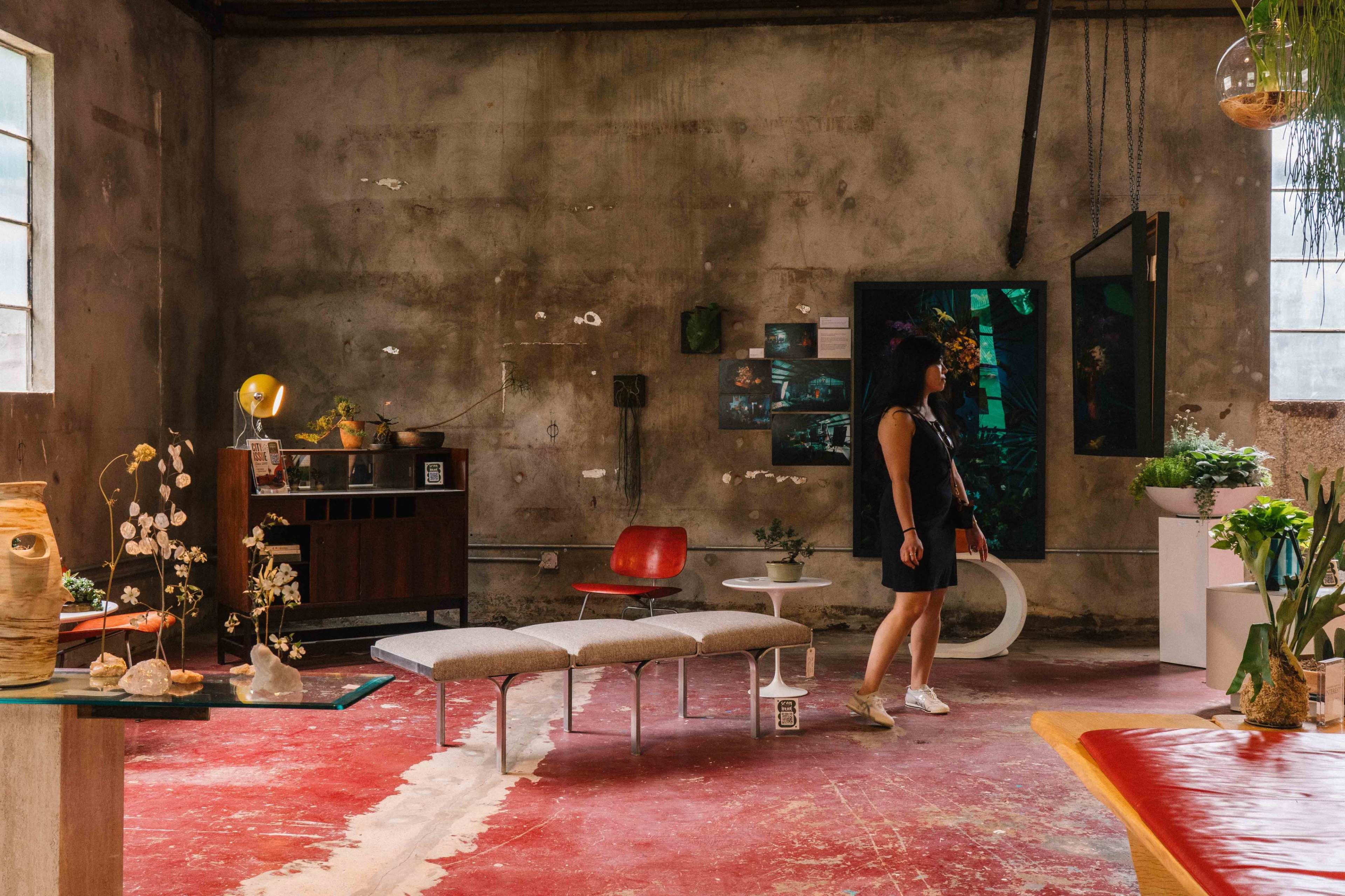 A woman walks through an artistic interior space featuring vintage furniture, plants, and various decorative items against a textured wall.