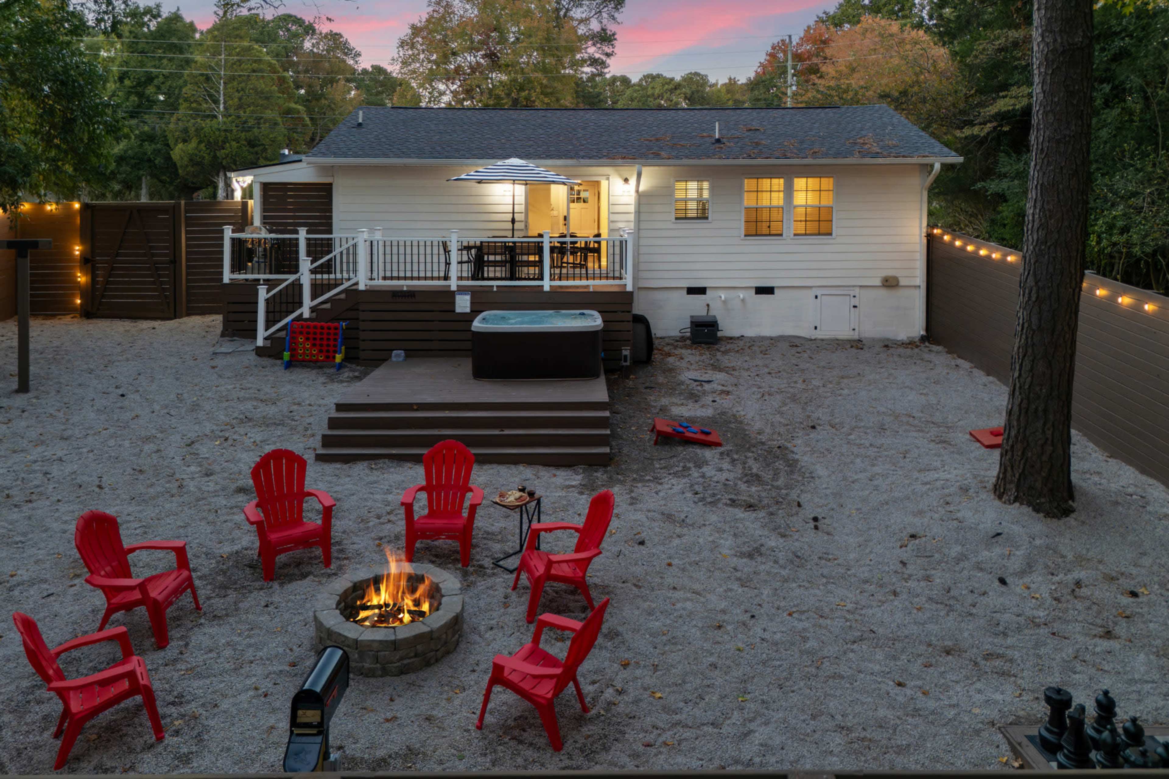 The image shows a cozy backyard with a fire pit surrounded by red chairs, a hot tub, and a house in the background, all set against a dusky sky.