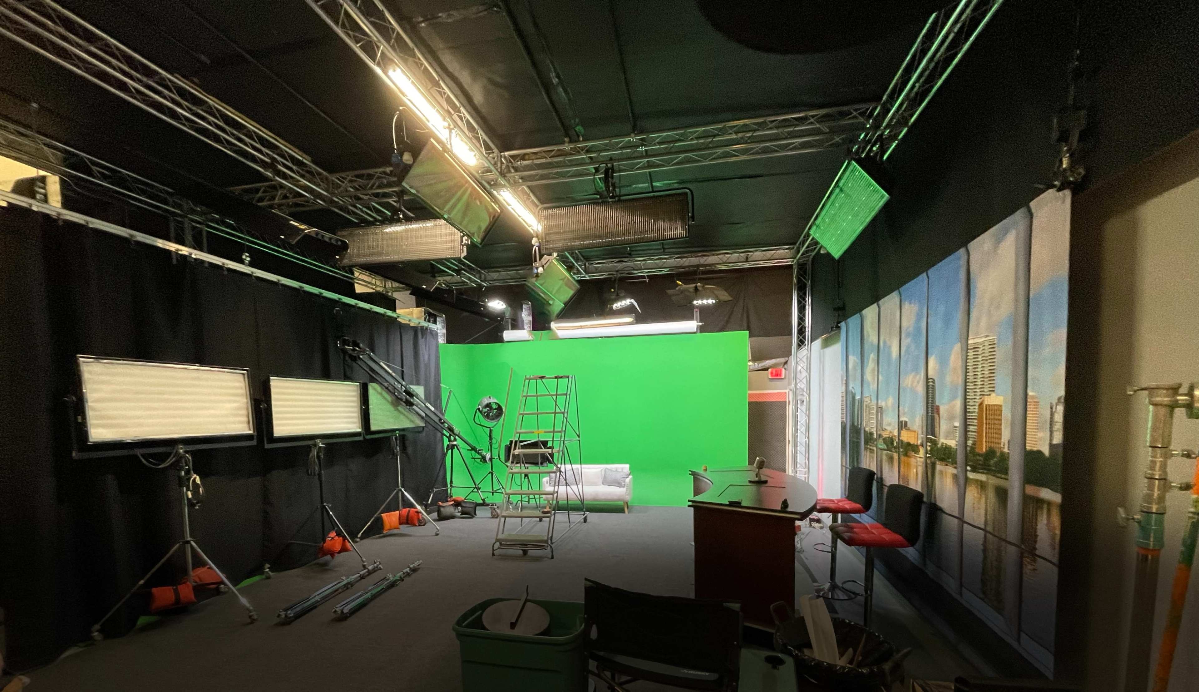 A television studio equipped with a green screen, lighting equipment, and a set area with a backdrop of a cityscape.