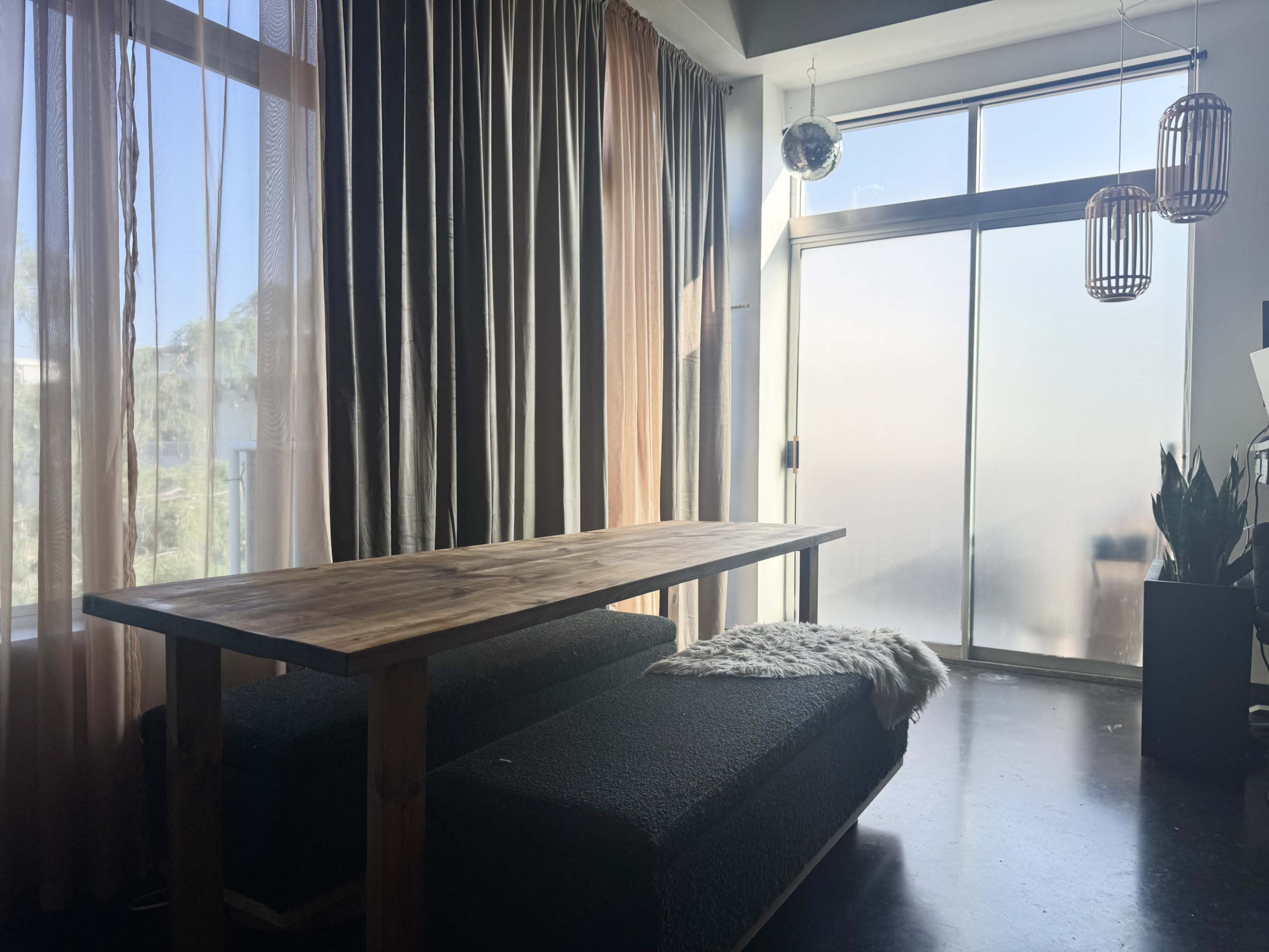 The image features a wooden table placed on a dark surface, next to a large window covered with sheer and solid curtains.