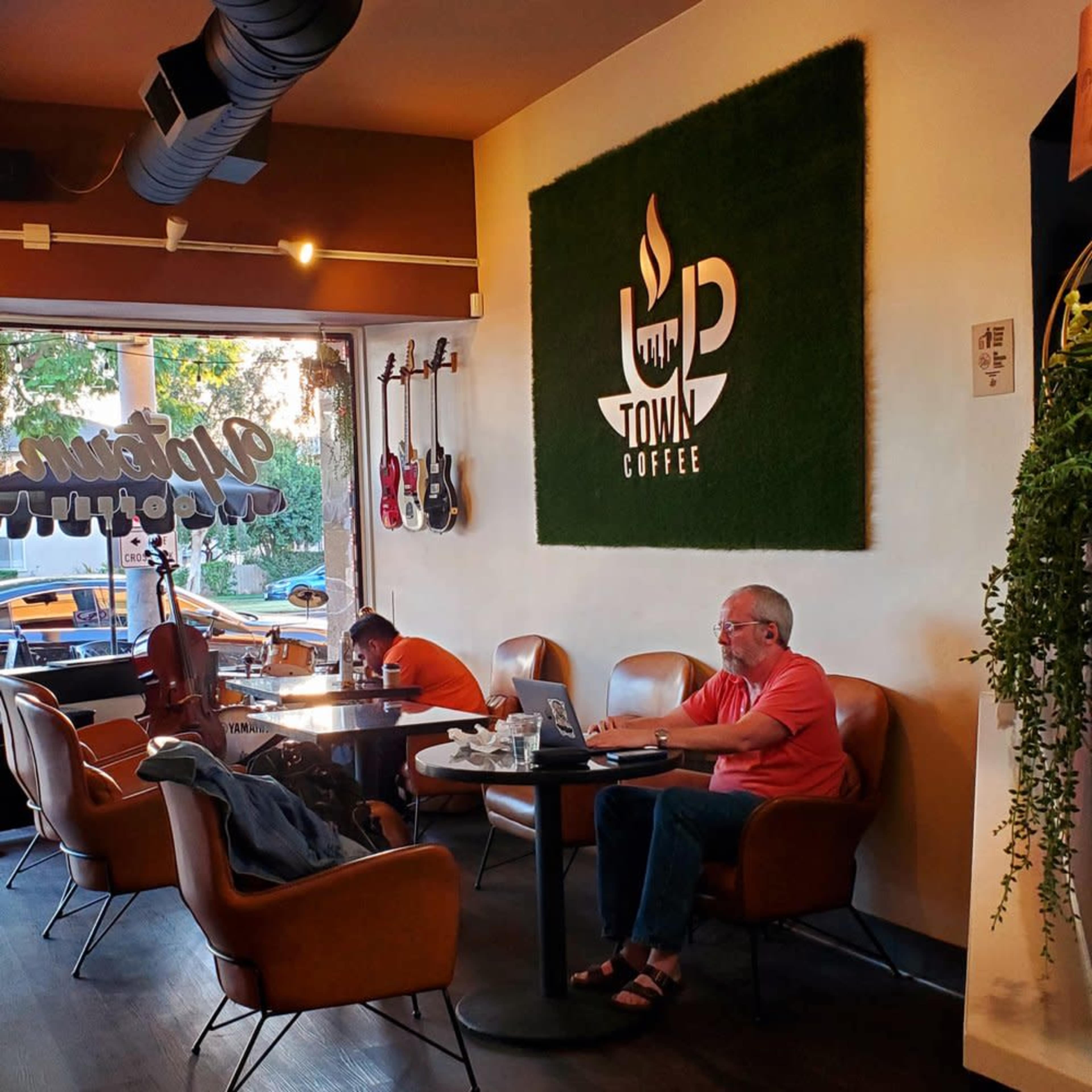 A coffee shop interior features two patrons, one working on a laptop and the other playing a cello, with a green wall logo displaying "Town Coffee."