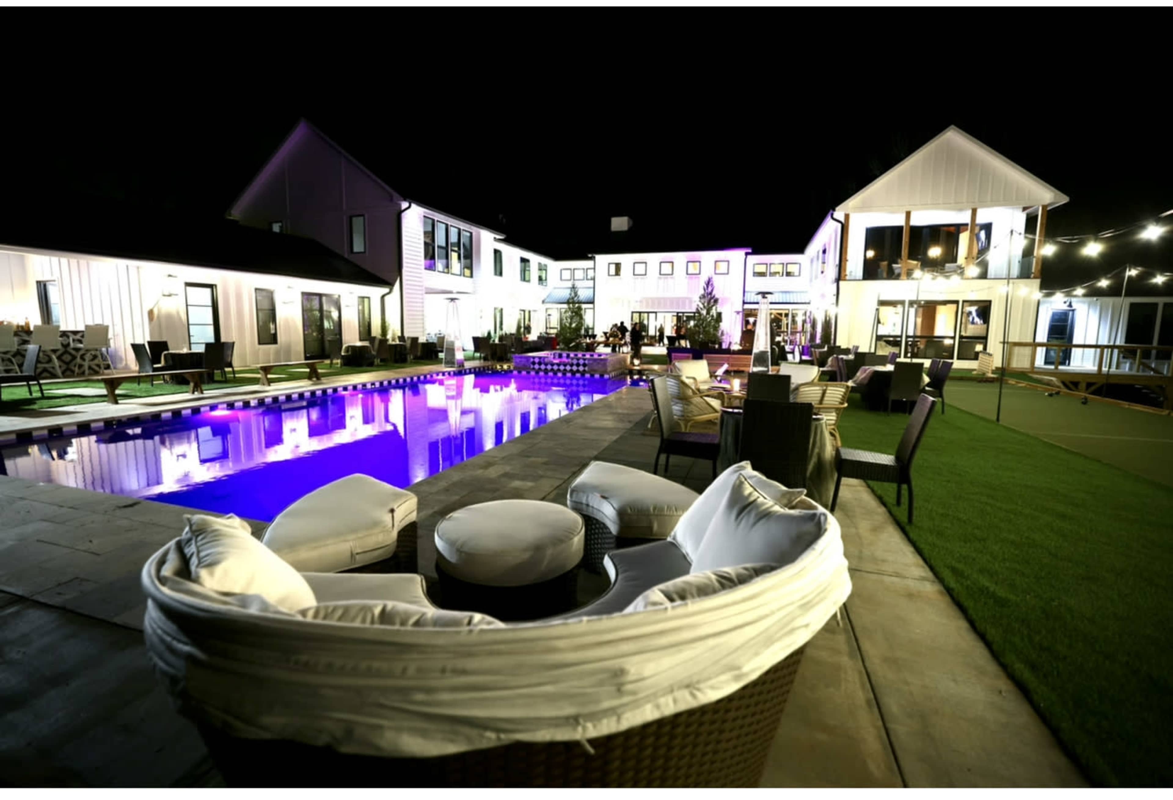 A modern outdoor space features a illuminated pool surrounded by lounge chairs and a spacious house with multiple levels, all set against a night sky.