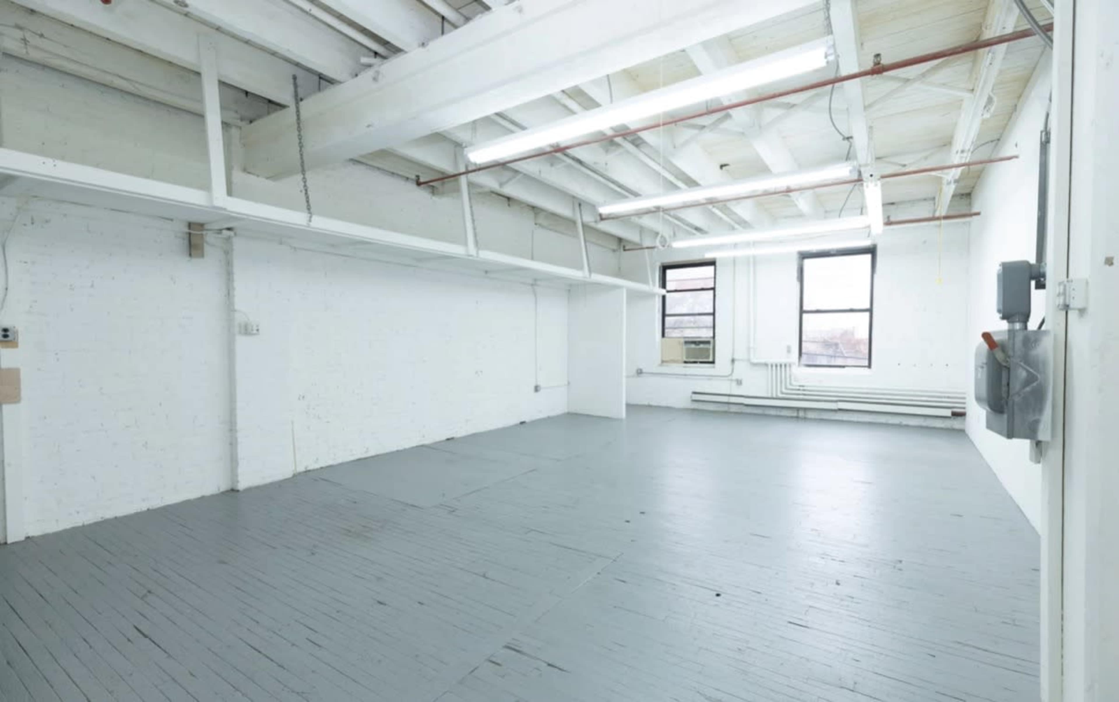 The image shows a spacious, empty room with high ceilings, wooden floors, and large windows letting in natural light.