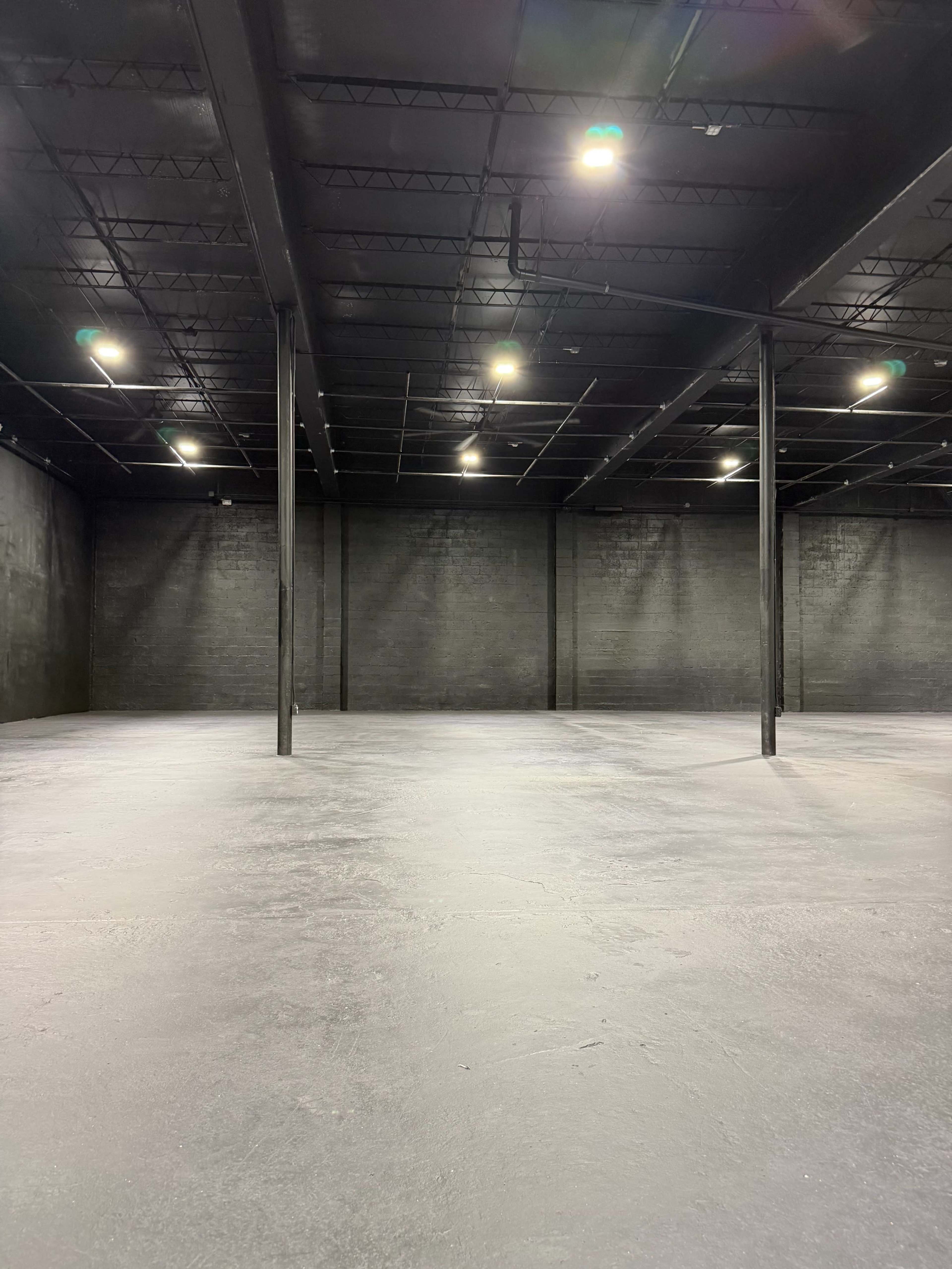 The image shows a large, empty warehouse interior with black walls and a smooth concrete floor, illuminated by overhead lights.