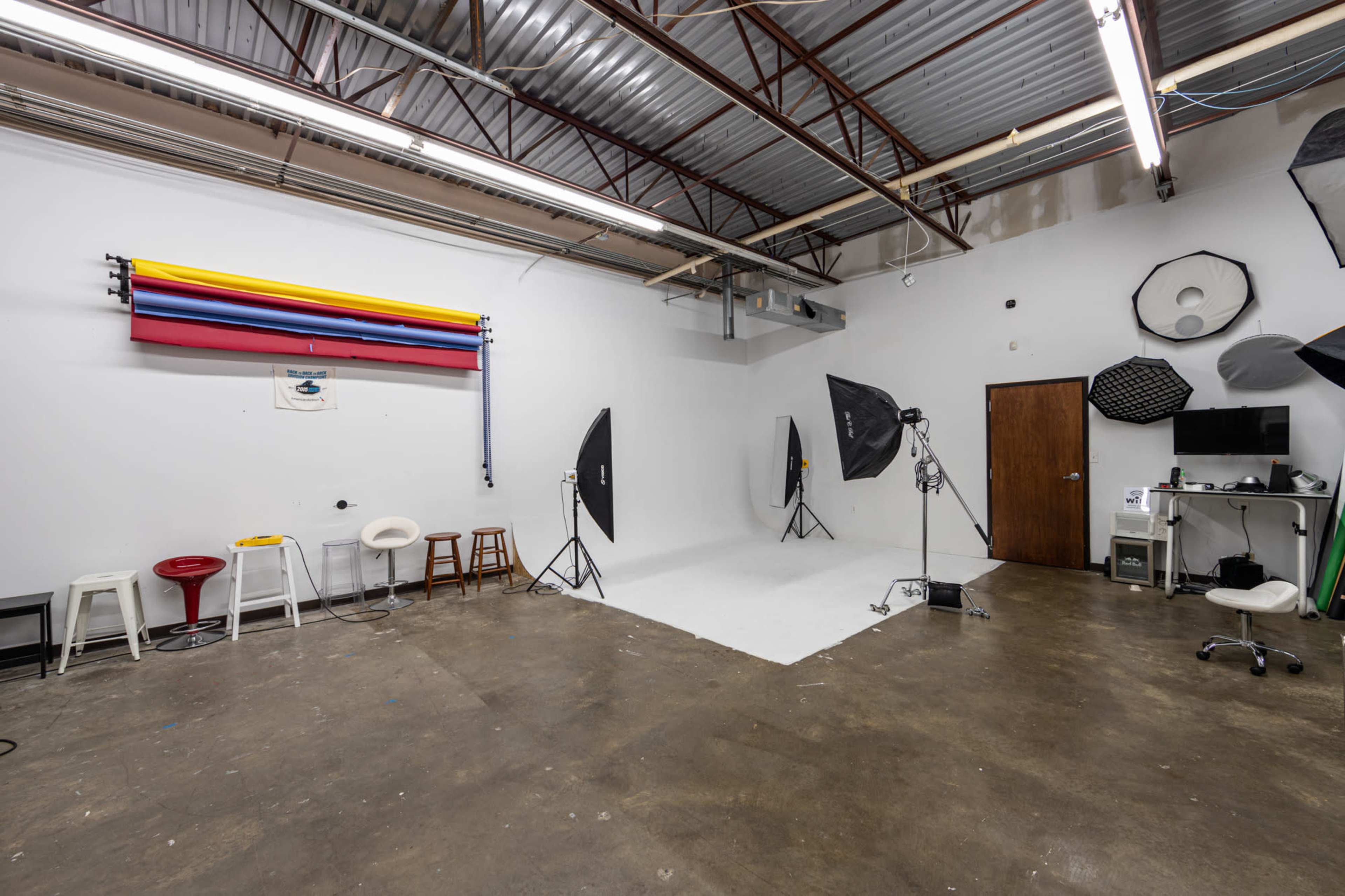 The image shows a photography studio with a white backdrop, lighting equipment, various stools, and a computer setup against a plain wall.