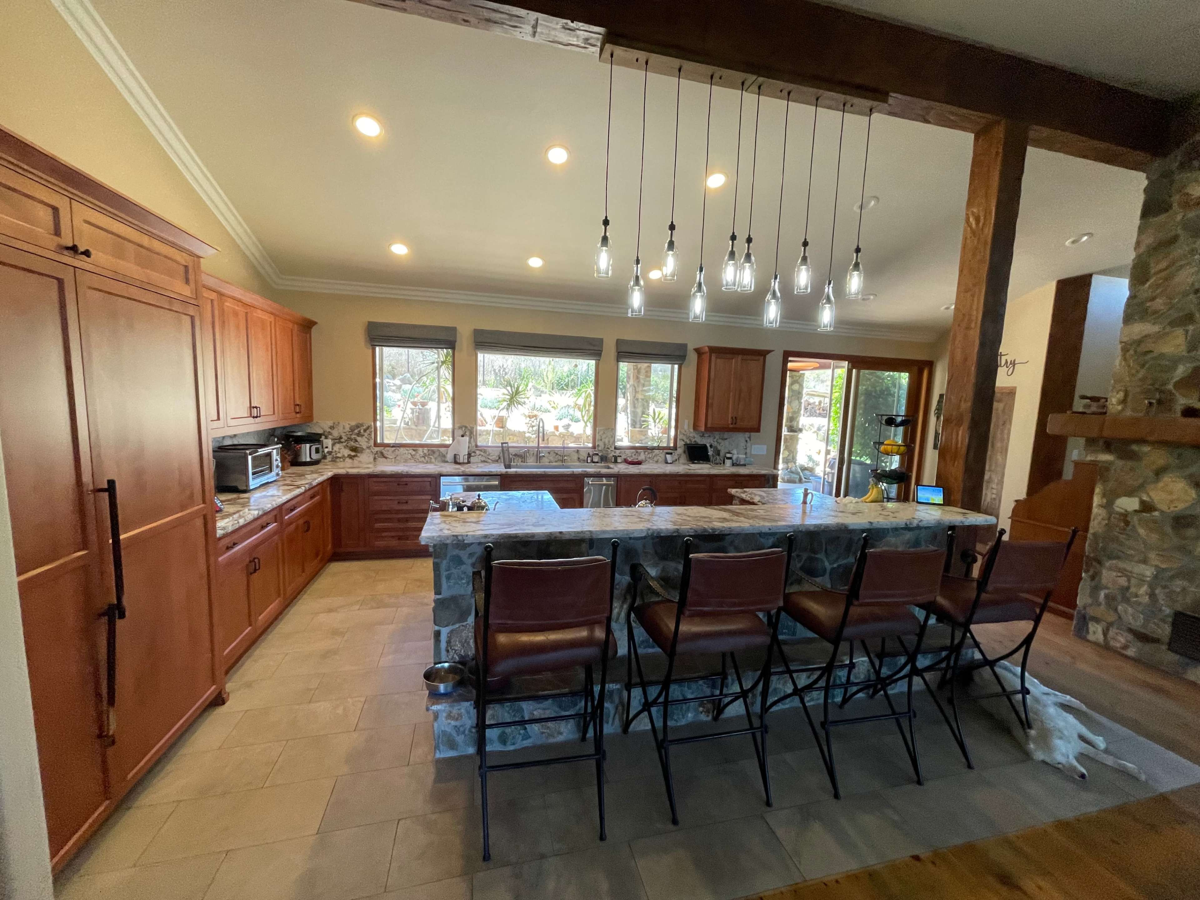The image shows a spacious kitchen with wooden cabinets, a large island with seating, and multiple windows letting in natural light.
