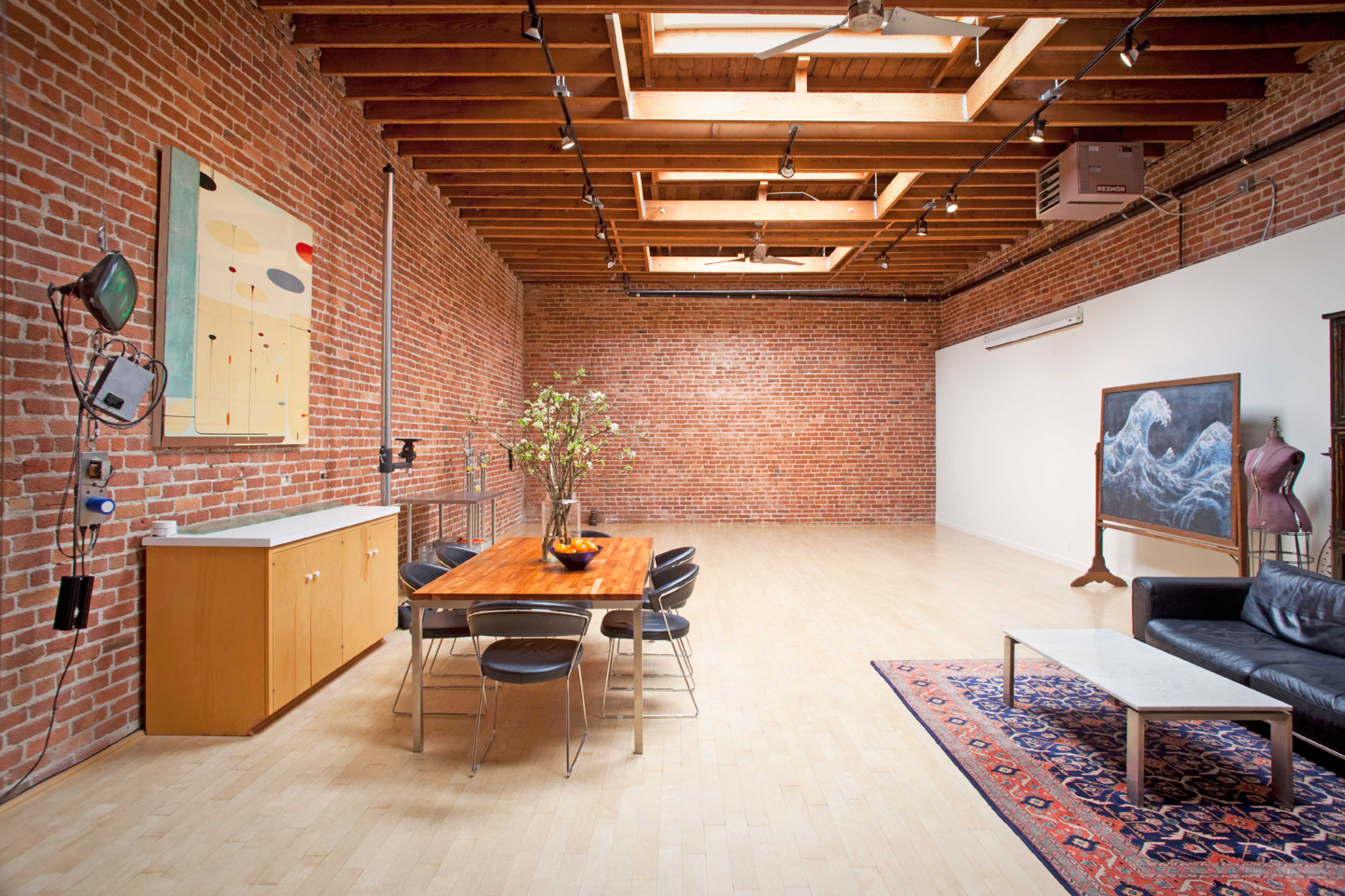 A spacious, industrial-style room with exposed brick walls, overhead beams, a wooden dining table surrounded by black chairs, a leather sofa, and an artwork display.