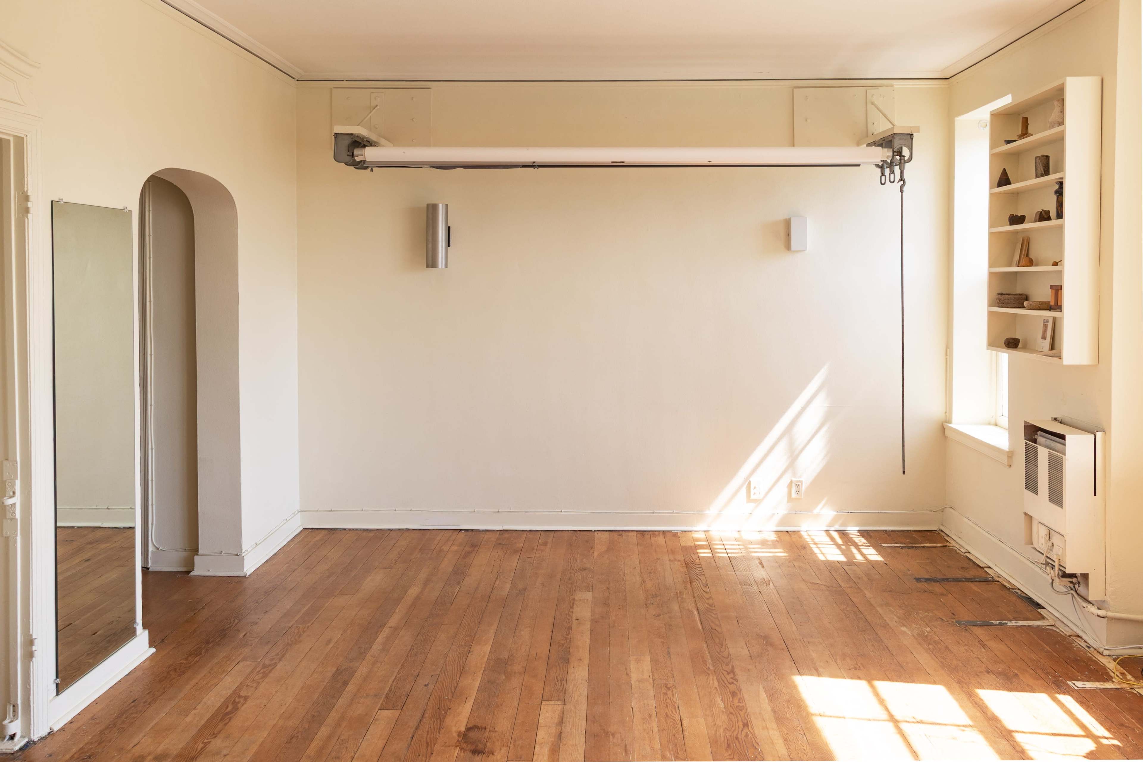 The image shows an empty room with wooden floors, a large mirror on one wall, a window with light streaming in, and minimal wall fixtures.
