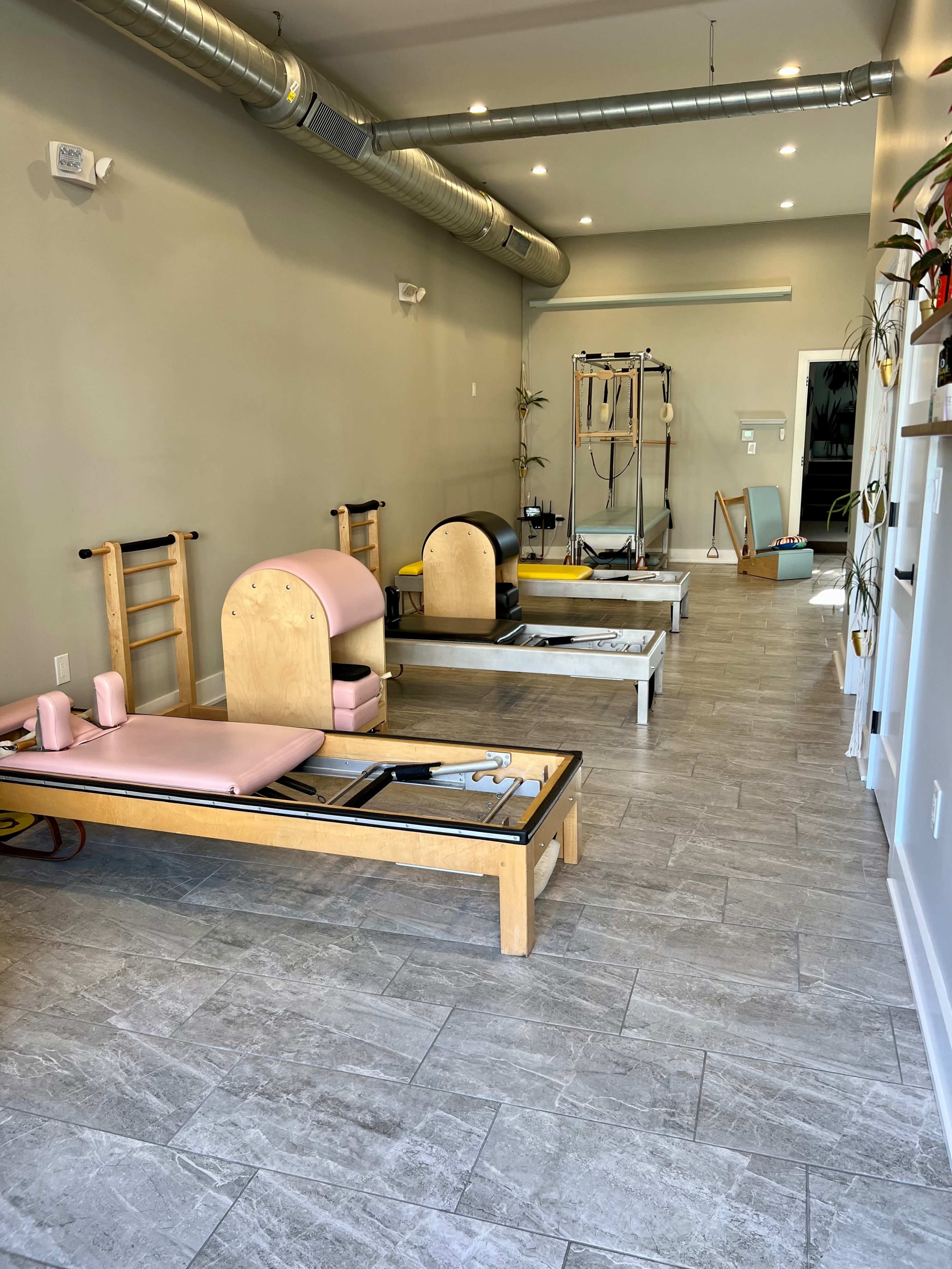 A modern Pilates studio featuring several reformer machines arranged along a spacious, light-colored floor with muted walls and overhead lighting.