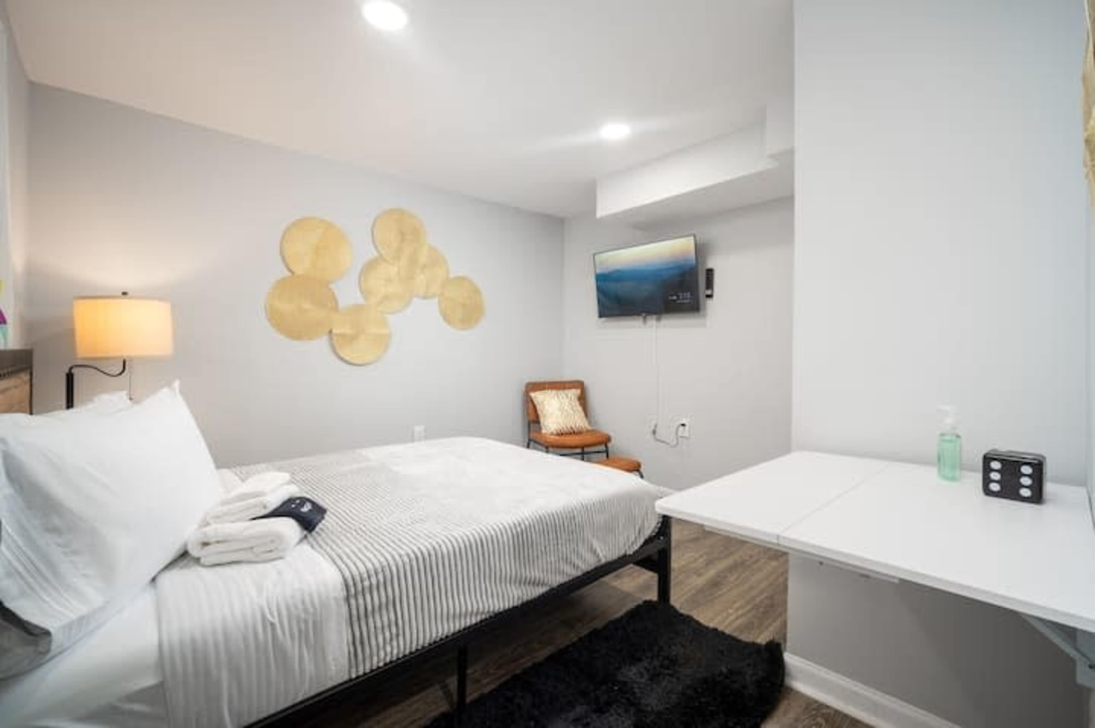A neatly arranged bedroom featuring a bed with white bedding, a small wooden chair, a wall-mounted TV, and a desk with hand sanitizer.