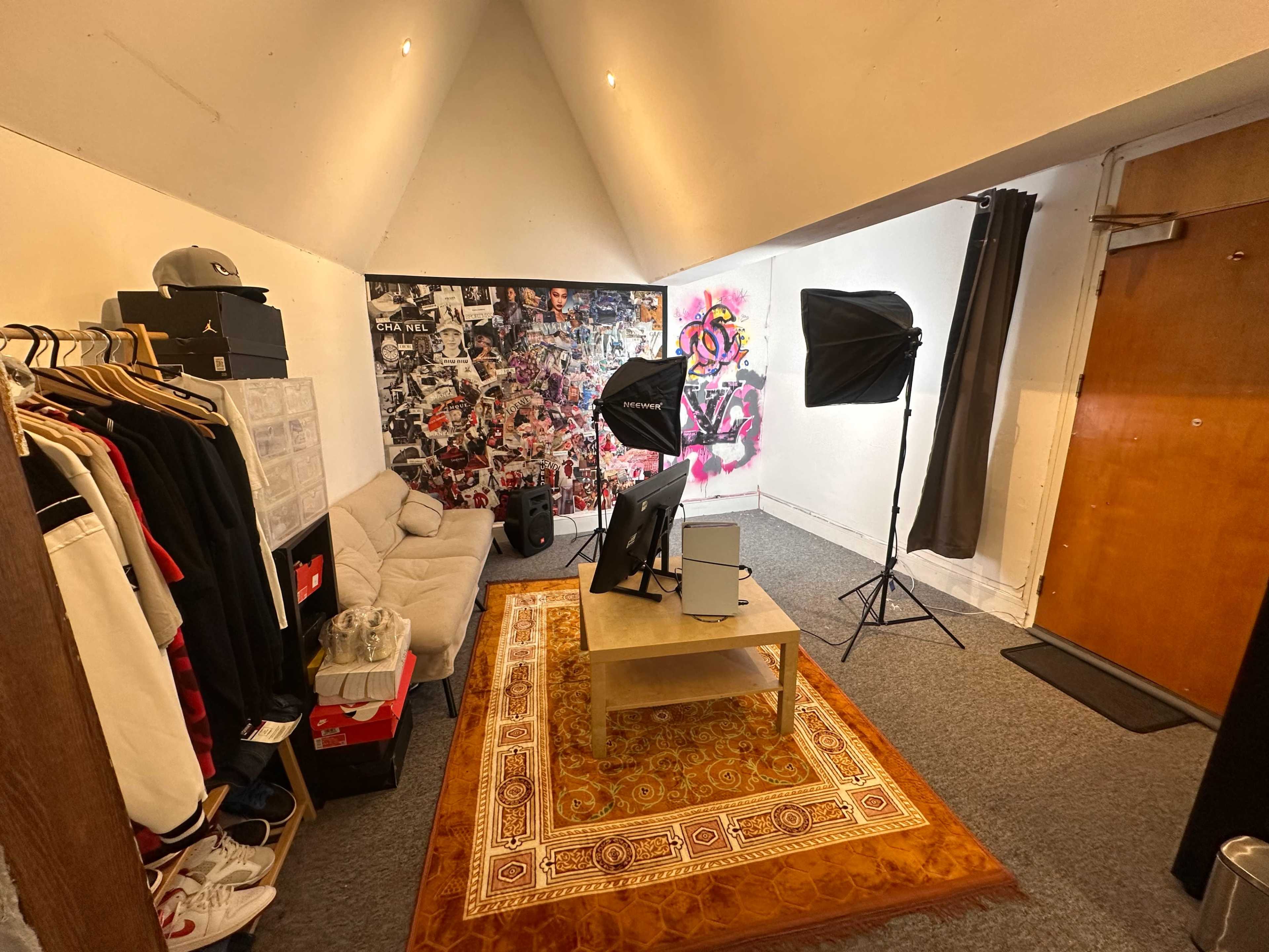 The image shows a small room with a couch, a coffee table, a television, photography lighting setups, and a wall covered in posters and artwork.