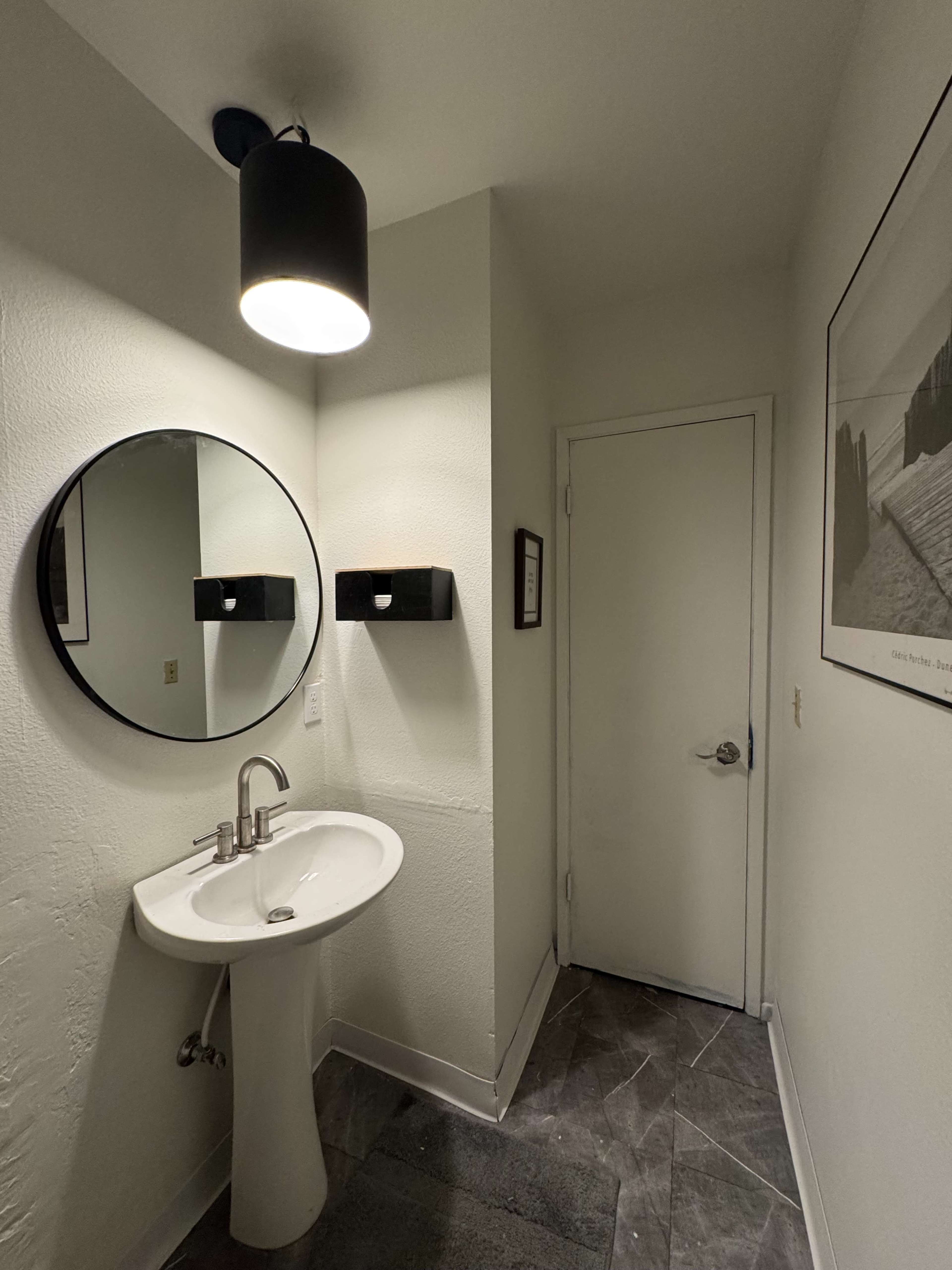 The image shows a small bathroom with a circular mirror above a pedestal sink, a light fixture on the ceiling, and a white door on the right.