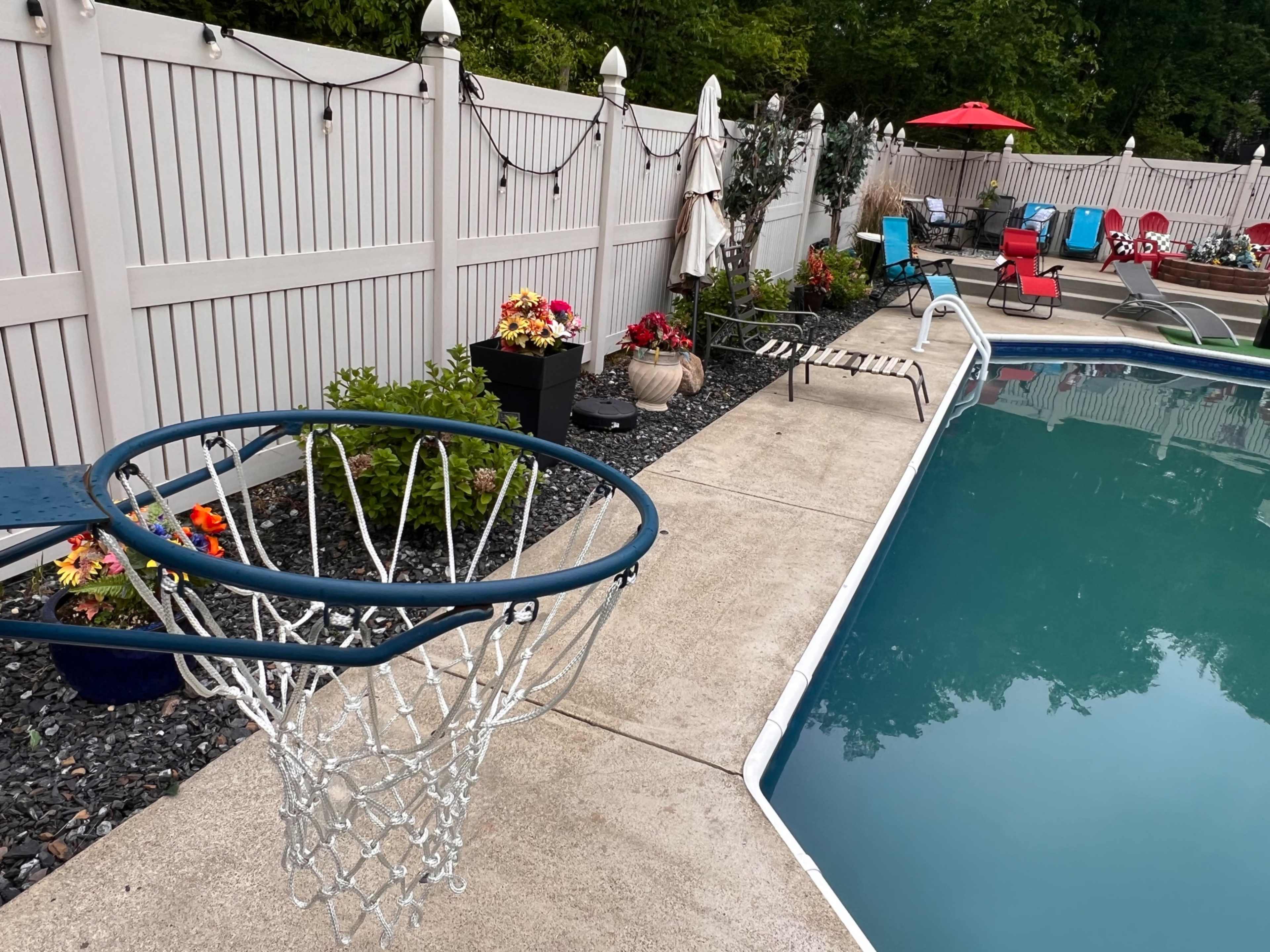 The image shows a basketball hoop next to a swimming pool, surrounded by decorative flowers and chairs.