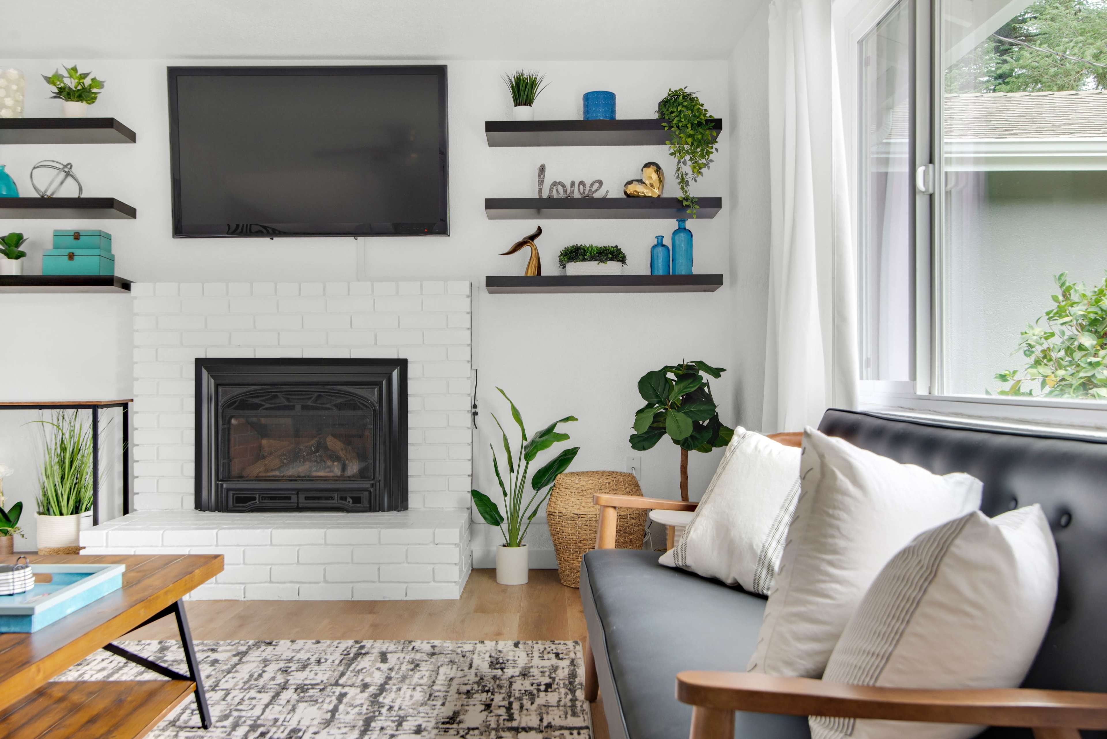 A living room features a black television mounted above a white brick fireplace, surrounded by shelves with decorative items, and a gray sofa with cushions, positioned next to a coffee table and plants.