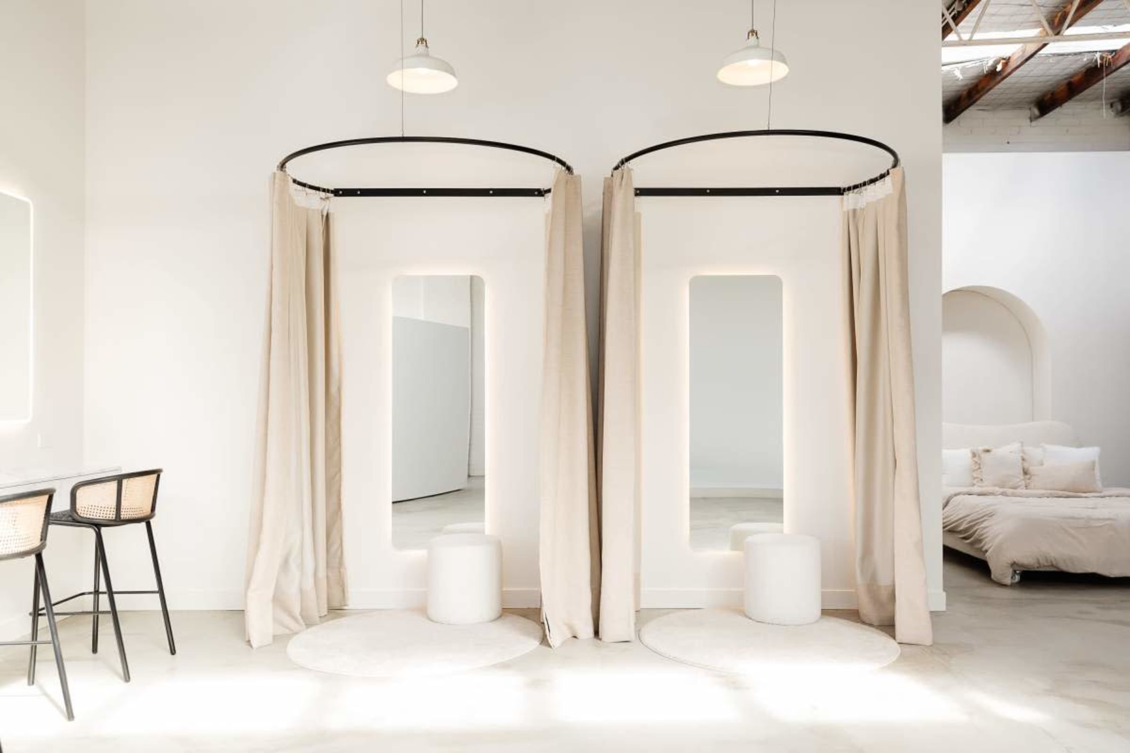 The image shows a minimalist interior with two dressing areas separated by soft curtains, each featuring a large mirror and a small stool, with a light-colored bed visible in the background.