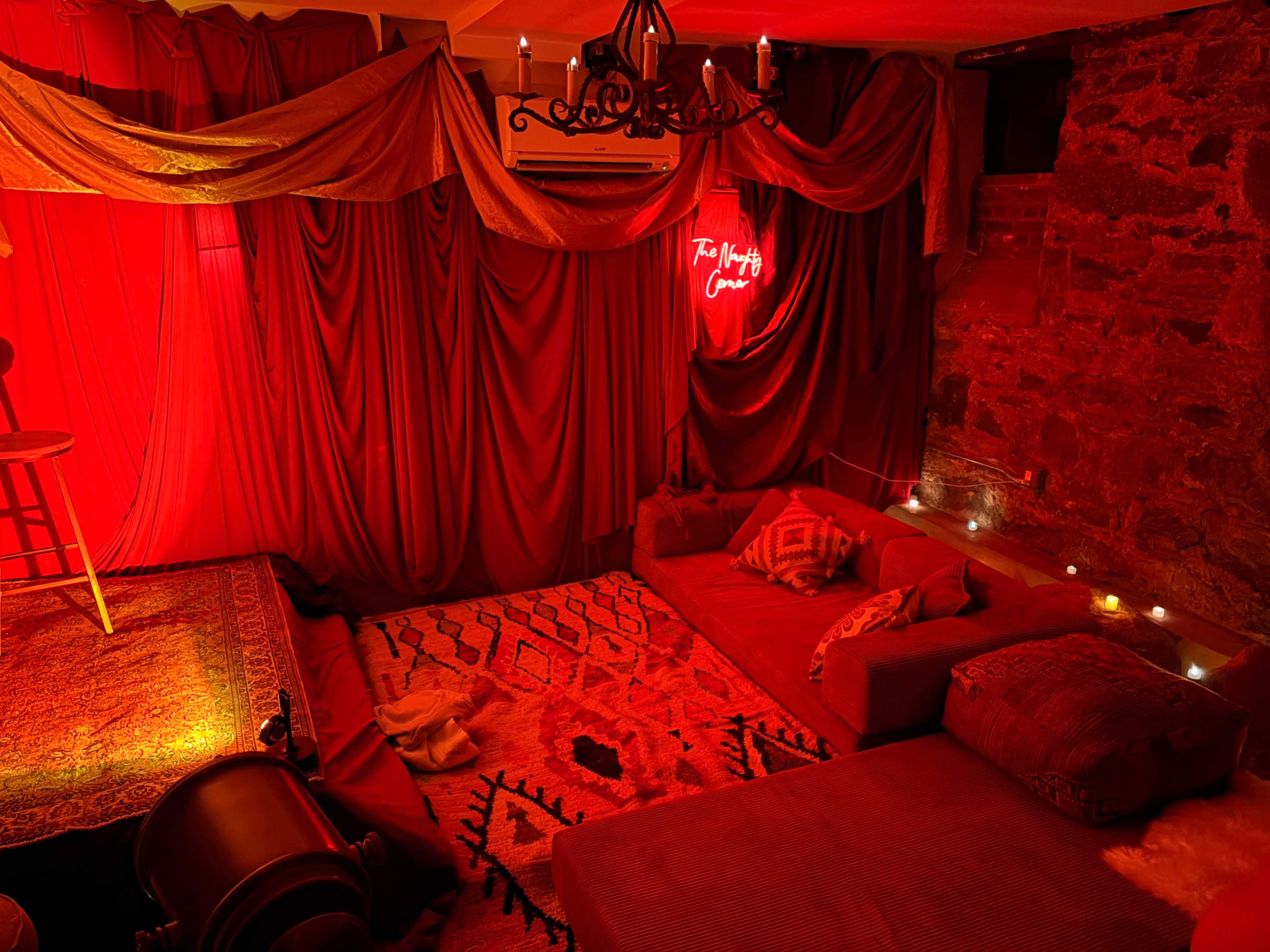 The image shows a cozy room illuminated with red lighting, featuring draped fabric, plush seating, and a neon sign that reads "The Night Cave."