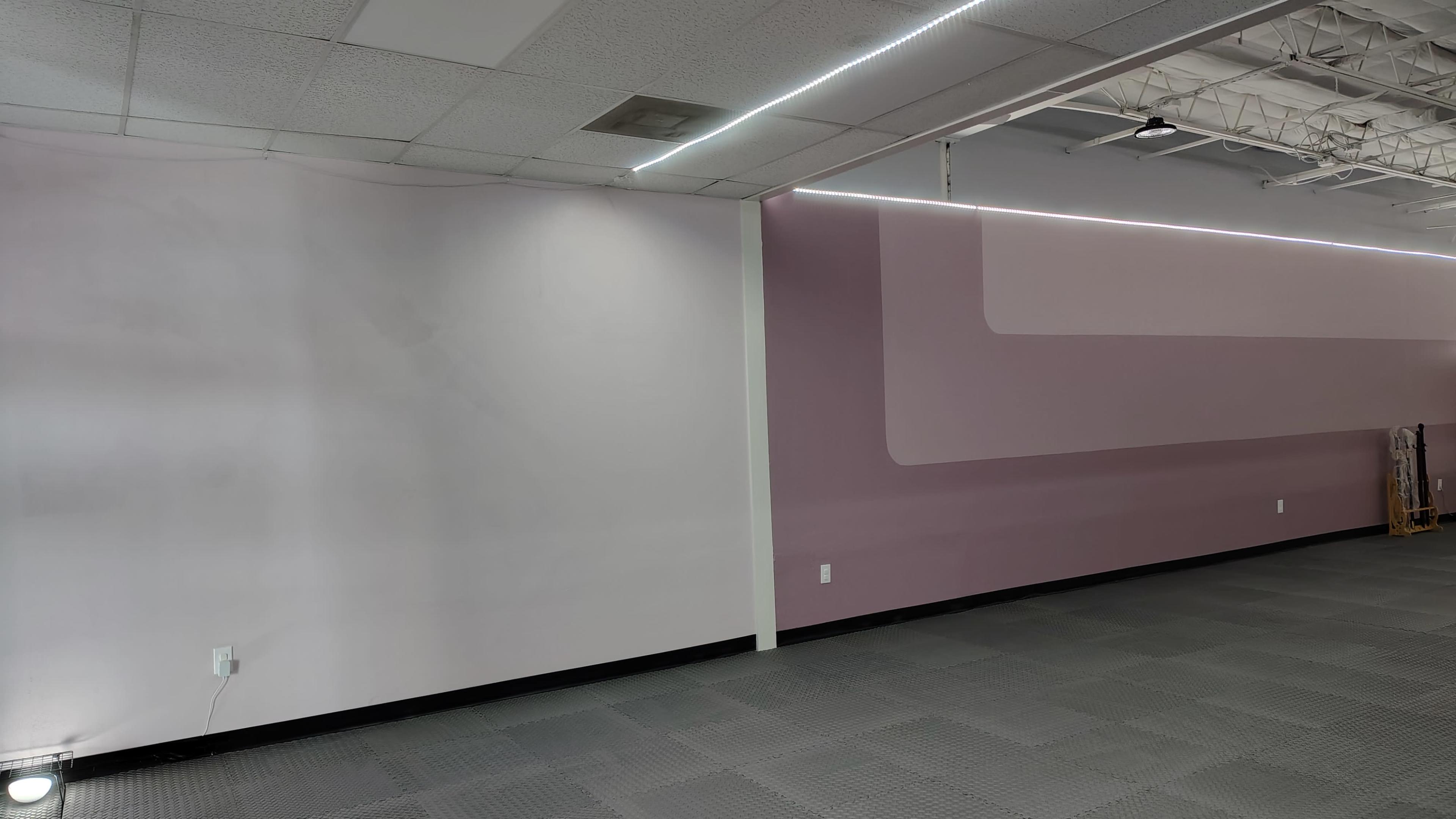 The image shows an empty interior space with light purple walls and gray flooring, featuring a section of wall with ambient lighting.