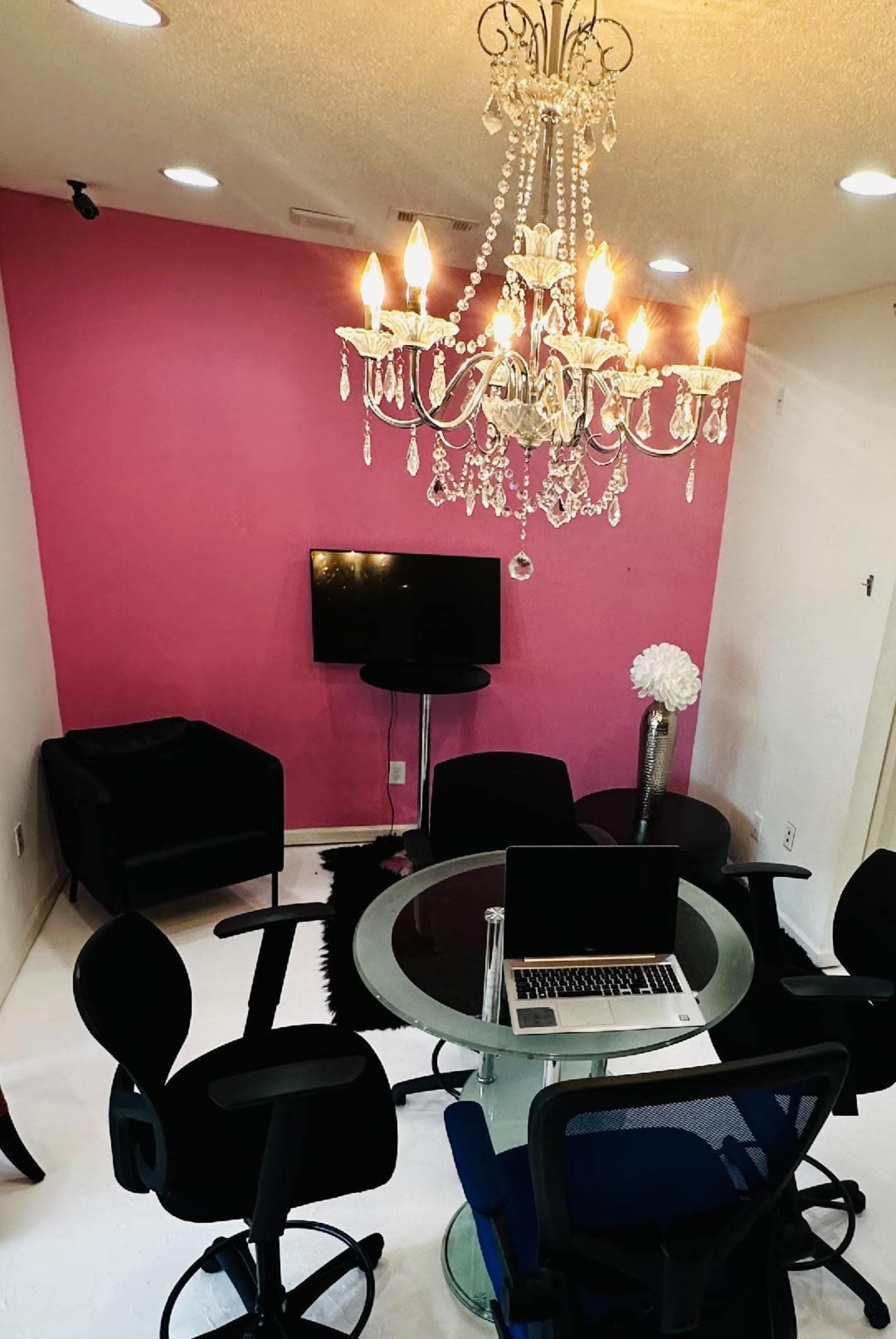 A small meeting room features a round glass table surrounded by black and blue office chairs, with a chandelier overhead and a pink accent wall.