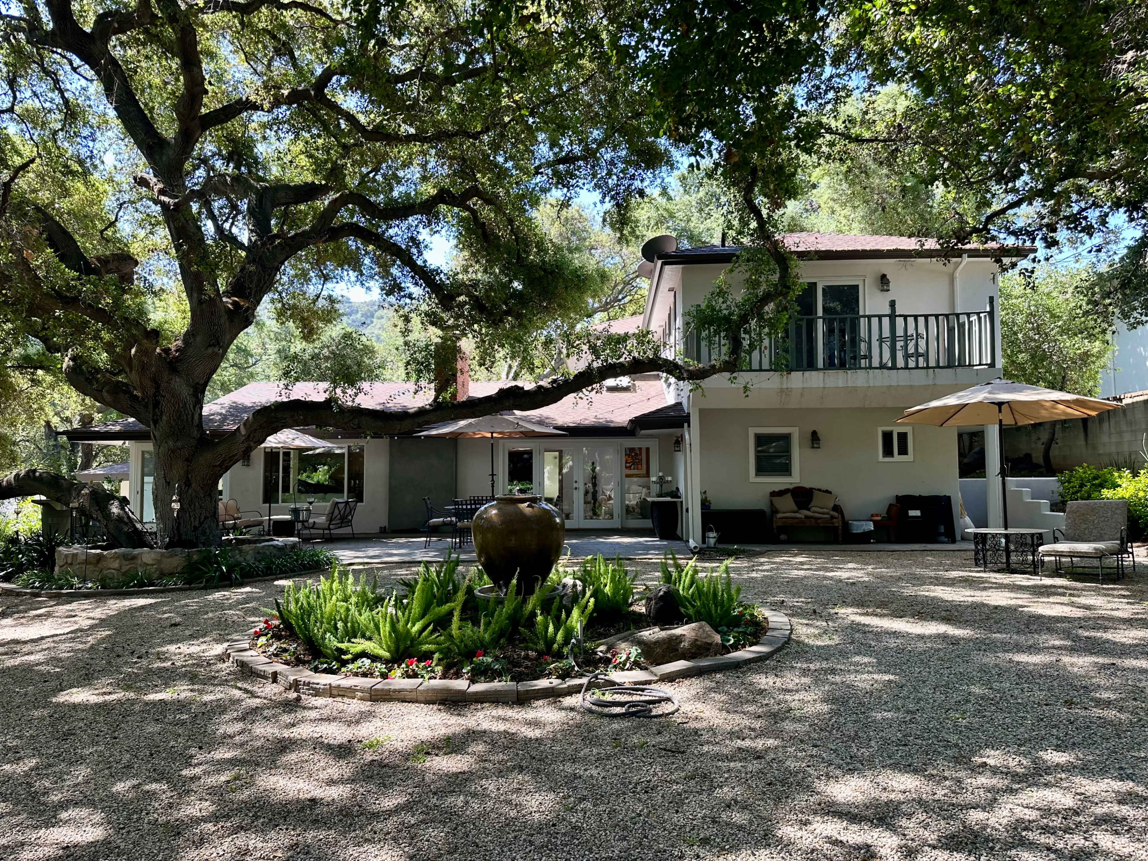A spacious house with a large tree in the front yard features a patio area and a decorative water fountain surrounded by landscaping.