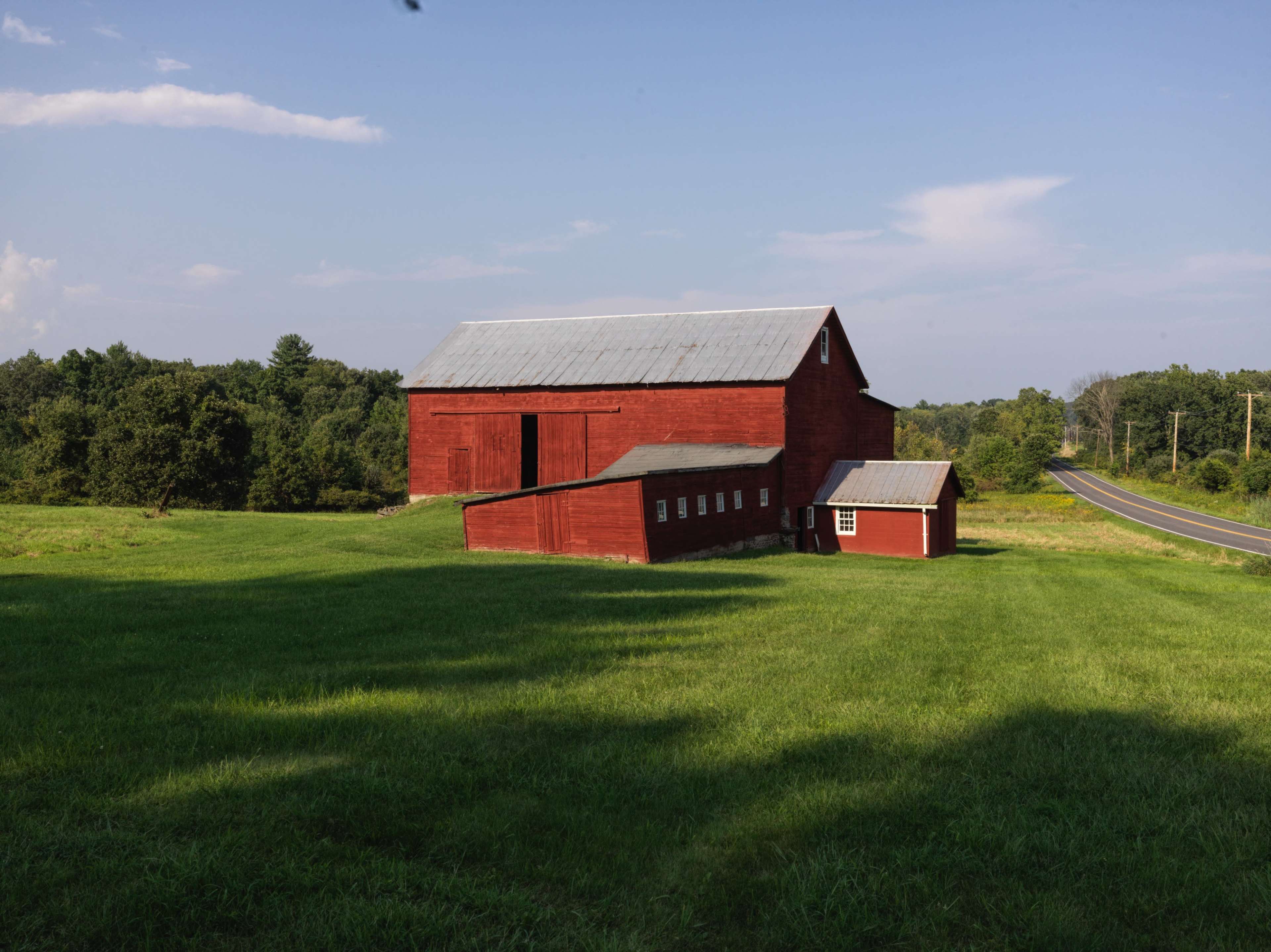 A large red barn with a metal roof sits on a grassy field next to a winding road.