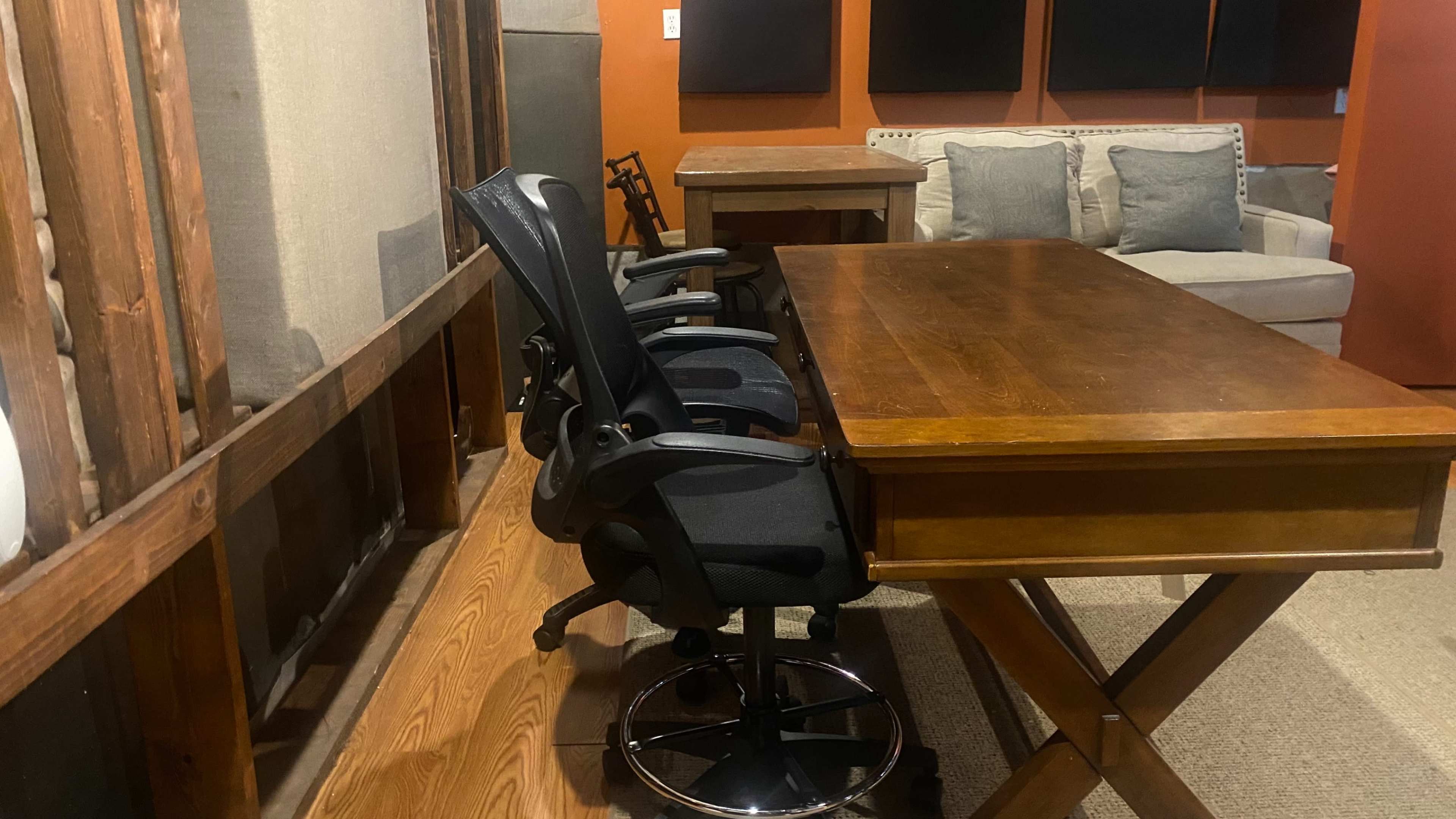 The image shows a wooden table with two office chairs positioned next to it, set in a room with exposed walls and a sofa in the background.