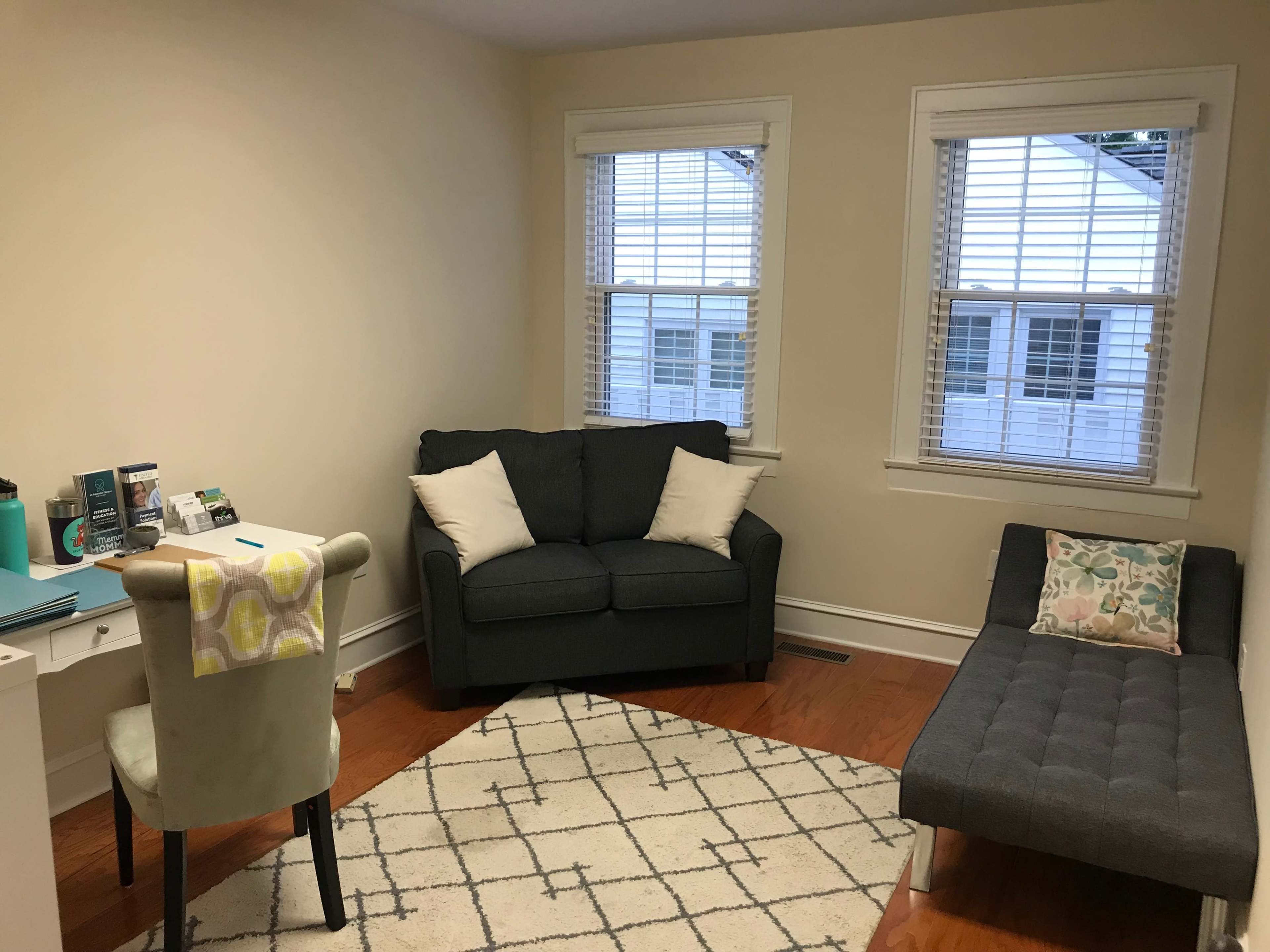 A small living room with a gray couch, a chaise lounge, a chair, and a desk next to a patterned rug and two windows.