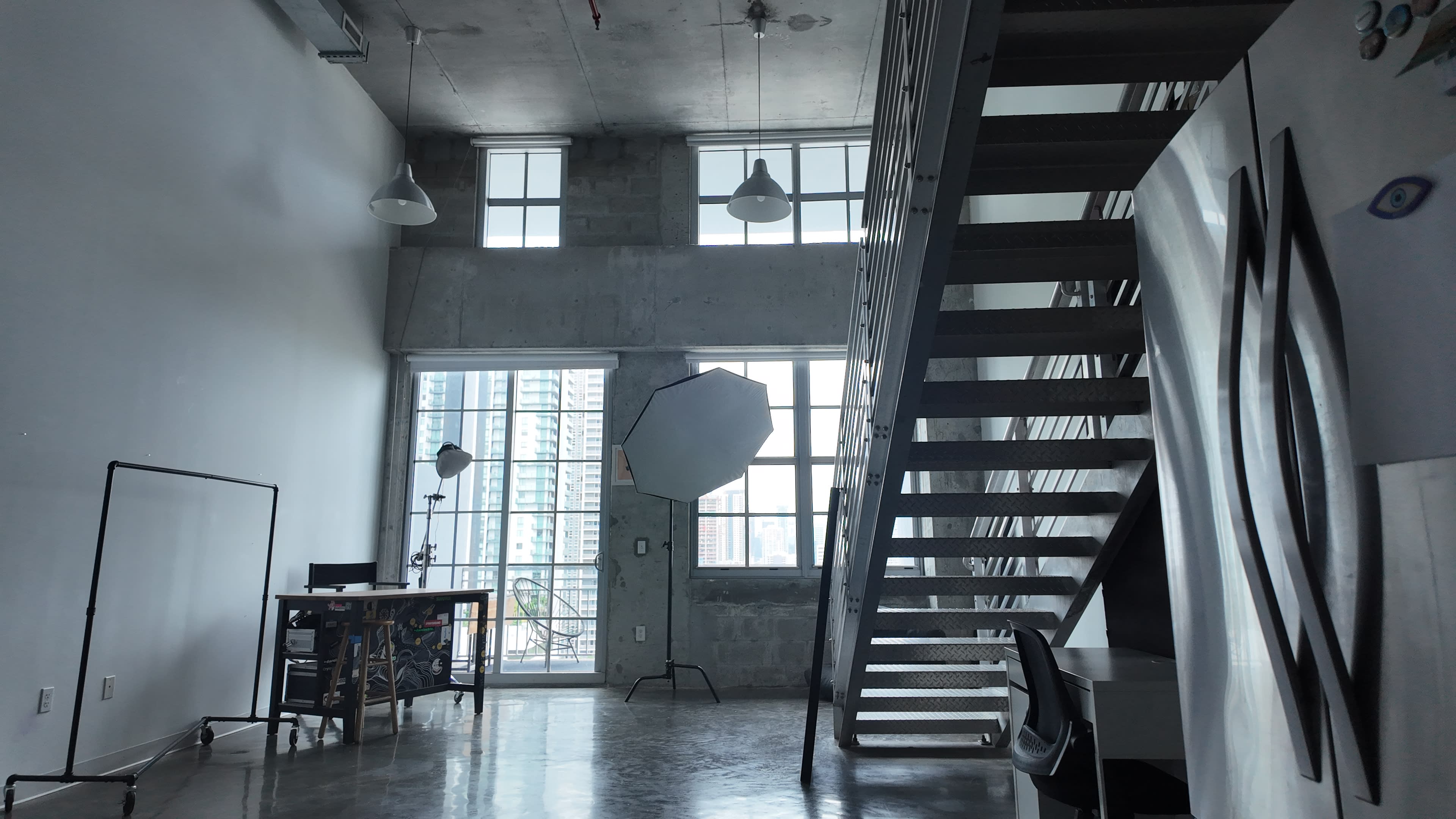 A loft-style room features large windows, a metal staircase, and a workspace with a desk and photography equipment.
