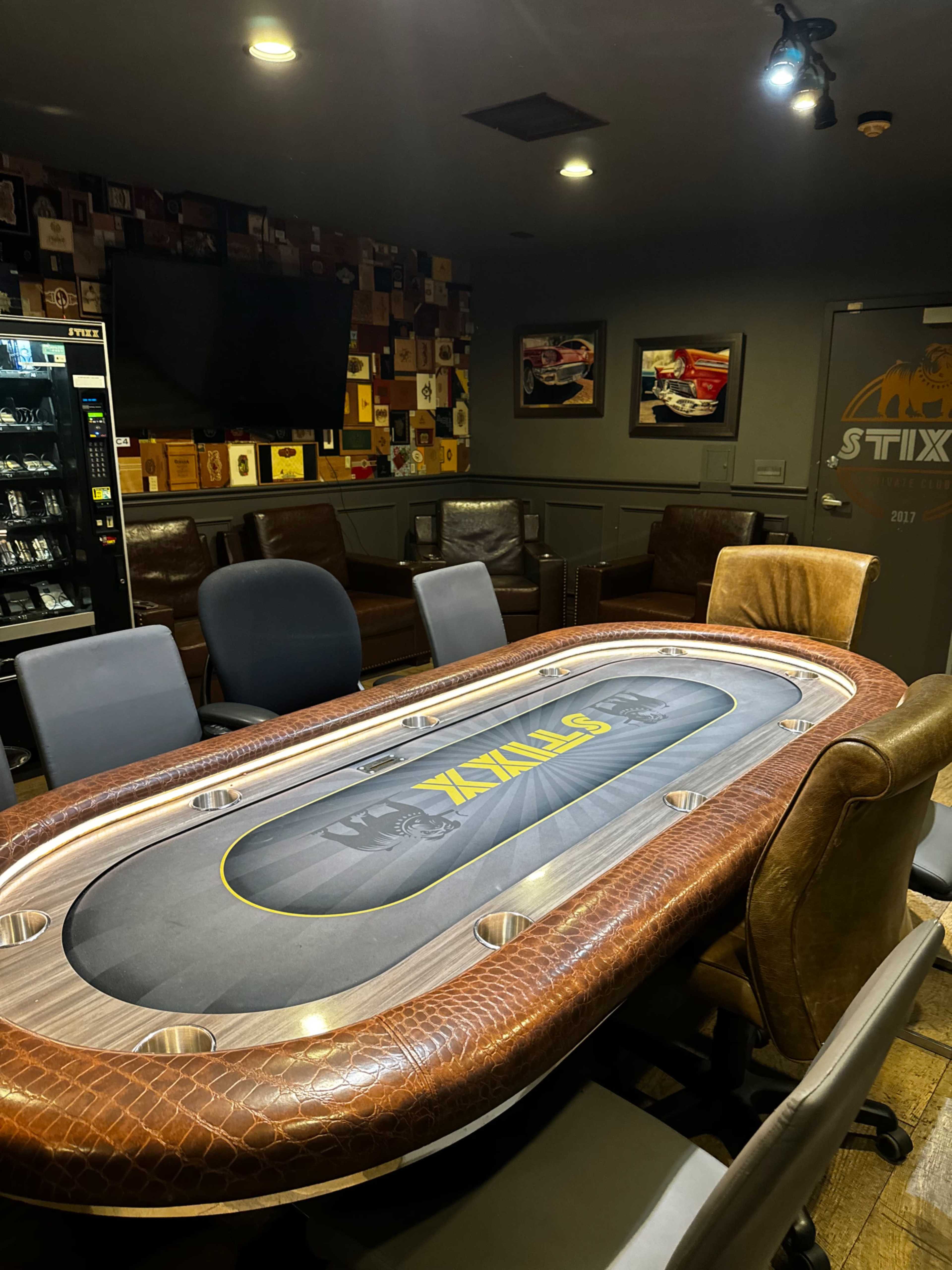 A poker table with a green felt surface is surrounded by various chairs in a dimly lit room decorated with framed pictures and a vending machine.