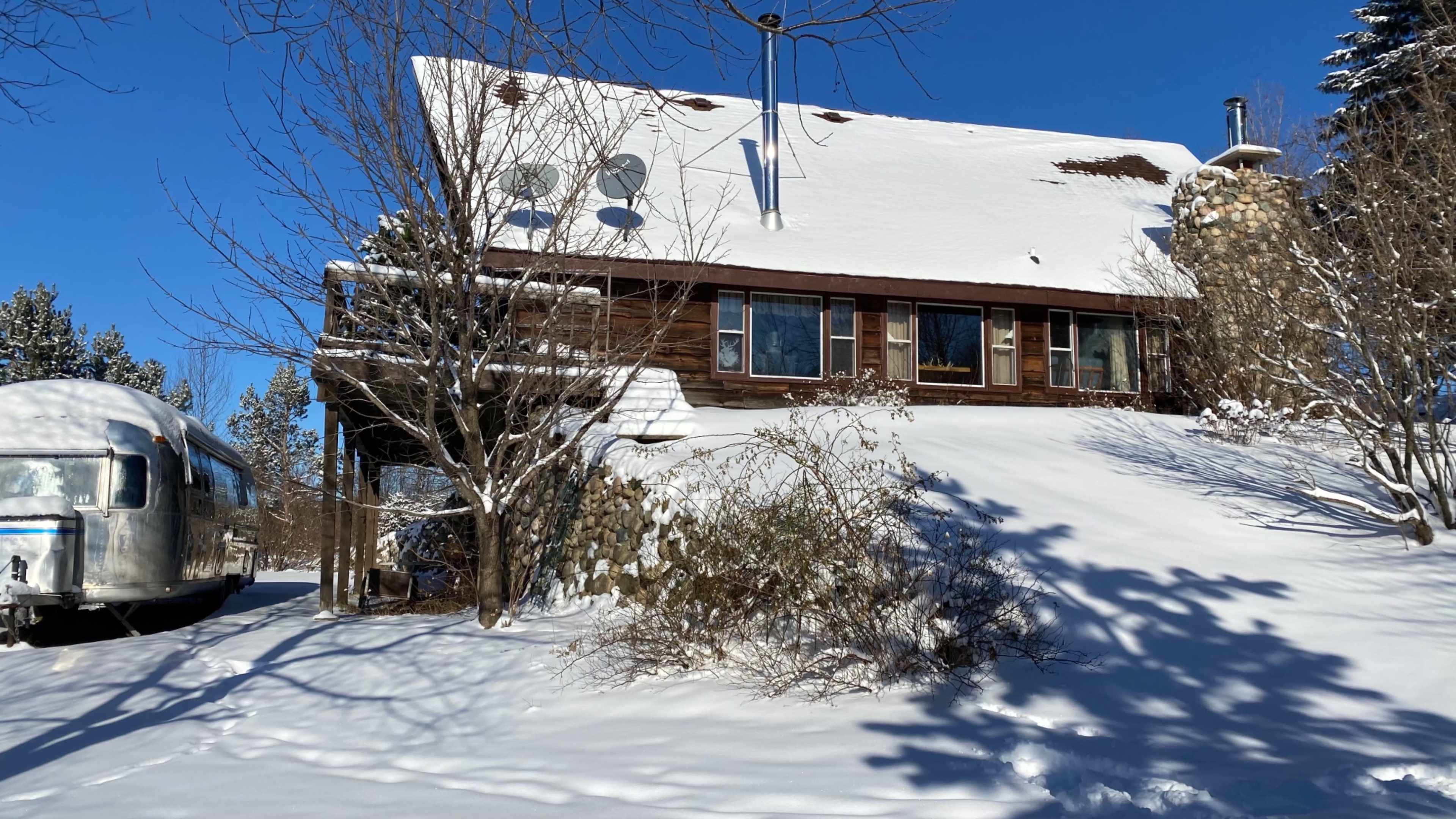 A wooden house with snow-covered roof and surrounding landscape is set on a hillside, partially obscured by trees and bushes, under a clear blue sky.