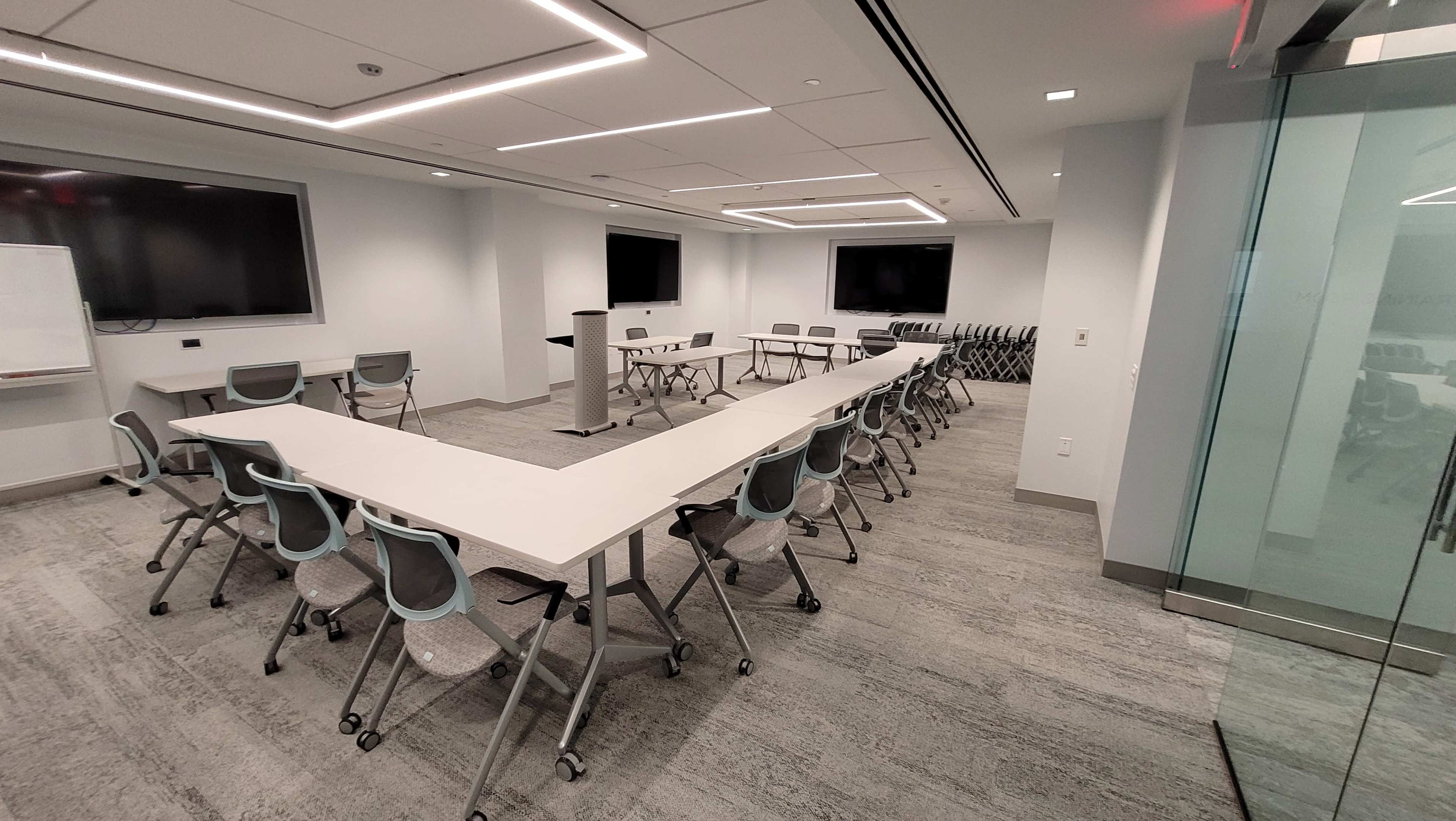 The image shows a modern meeting room arranged with a large U-shaped table surrounded by chairs, equipped with two large screens and a whiteboard.