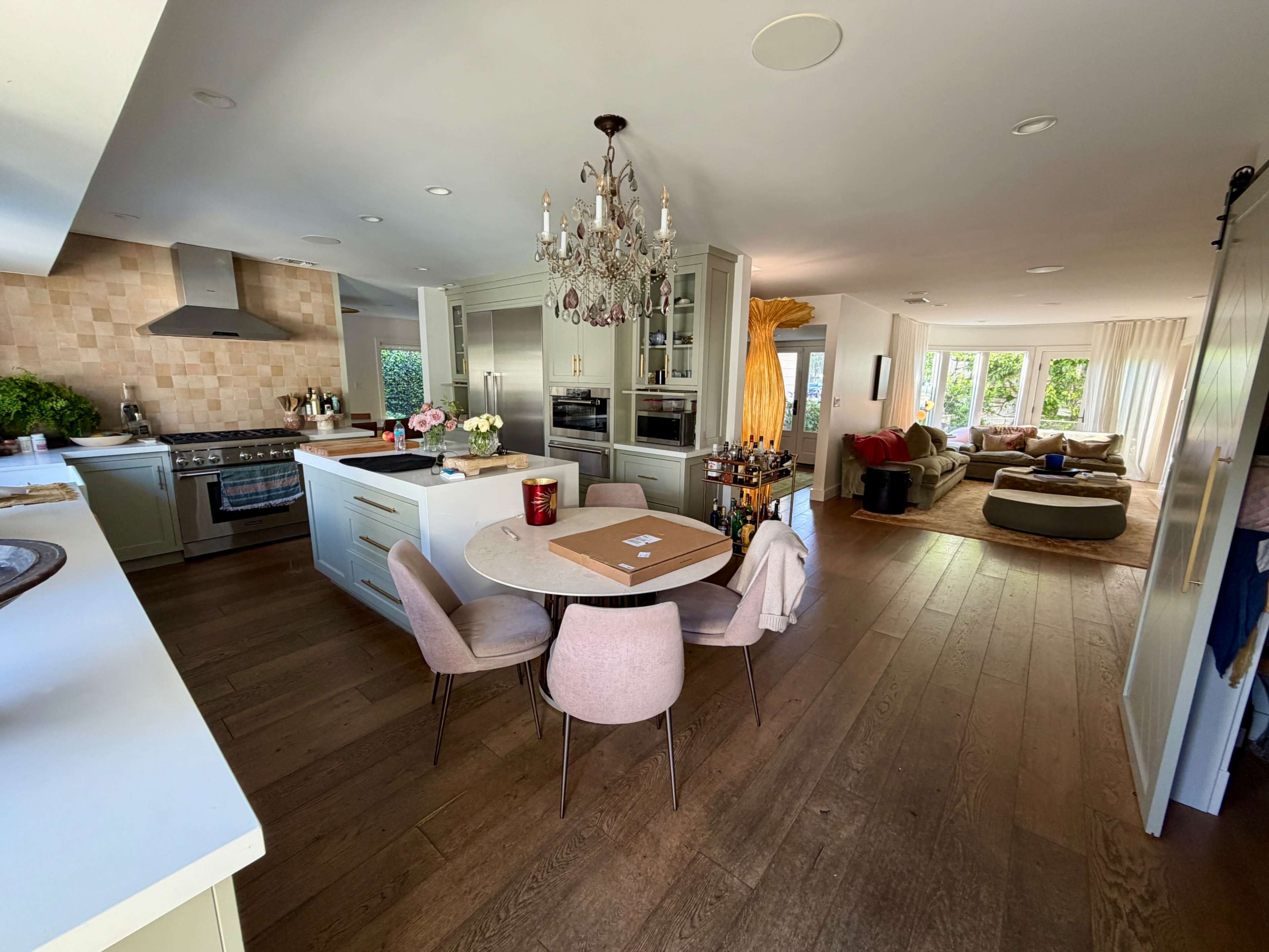 The image shows a modern kitchen and dining area featuring a round table with four chairs, a chandelier, and a spacious living room with a sectional sofa.