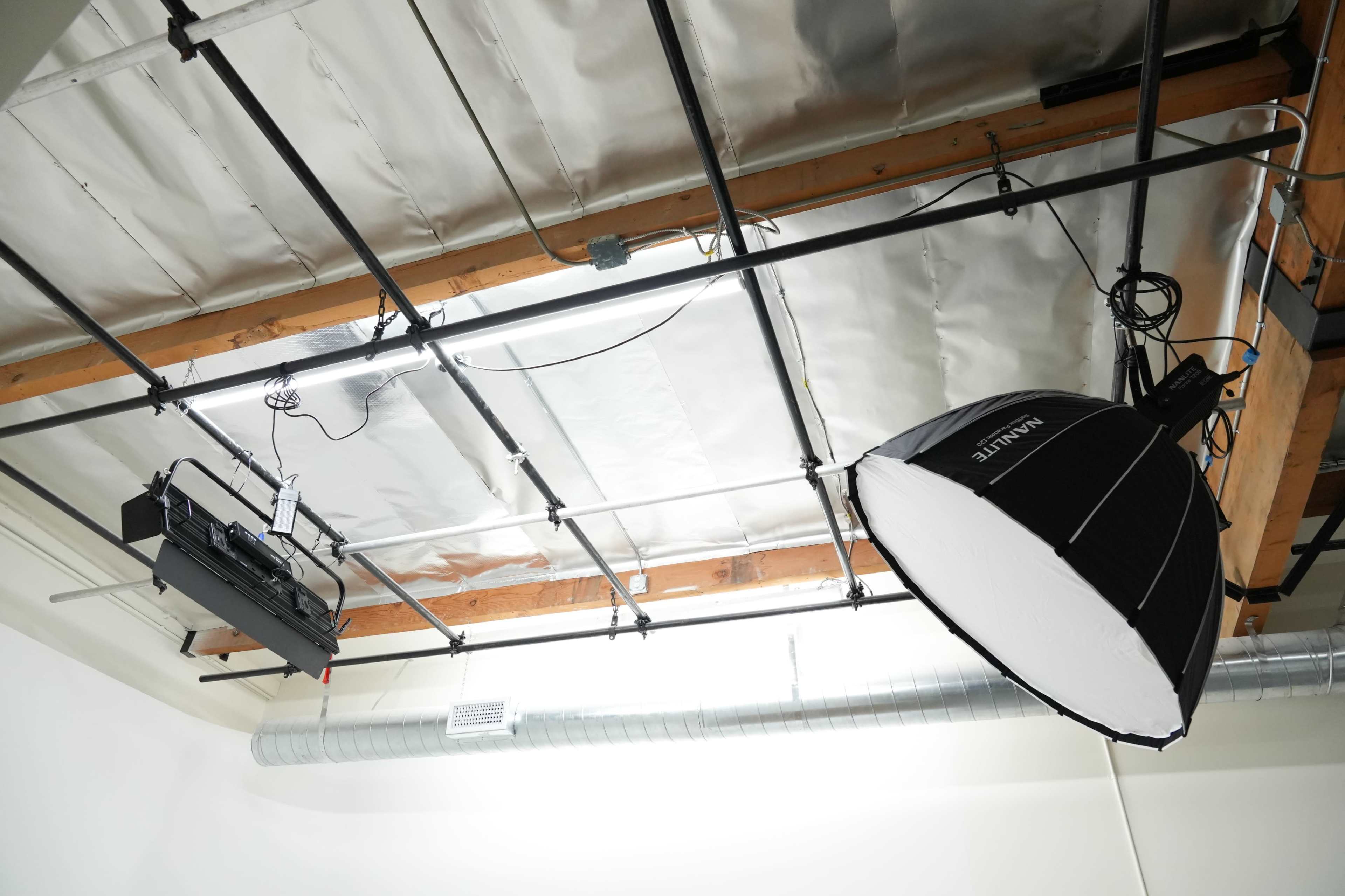 The image shows an overhead view of a studio space with exposed wooden beams, metal ducts, and lighting equipment, including a large softbox.