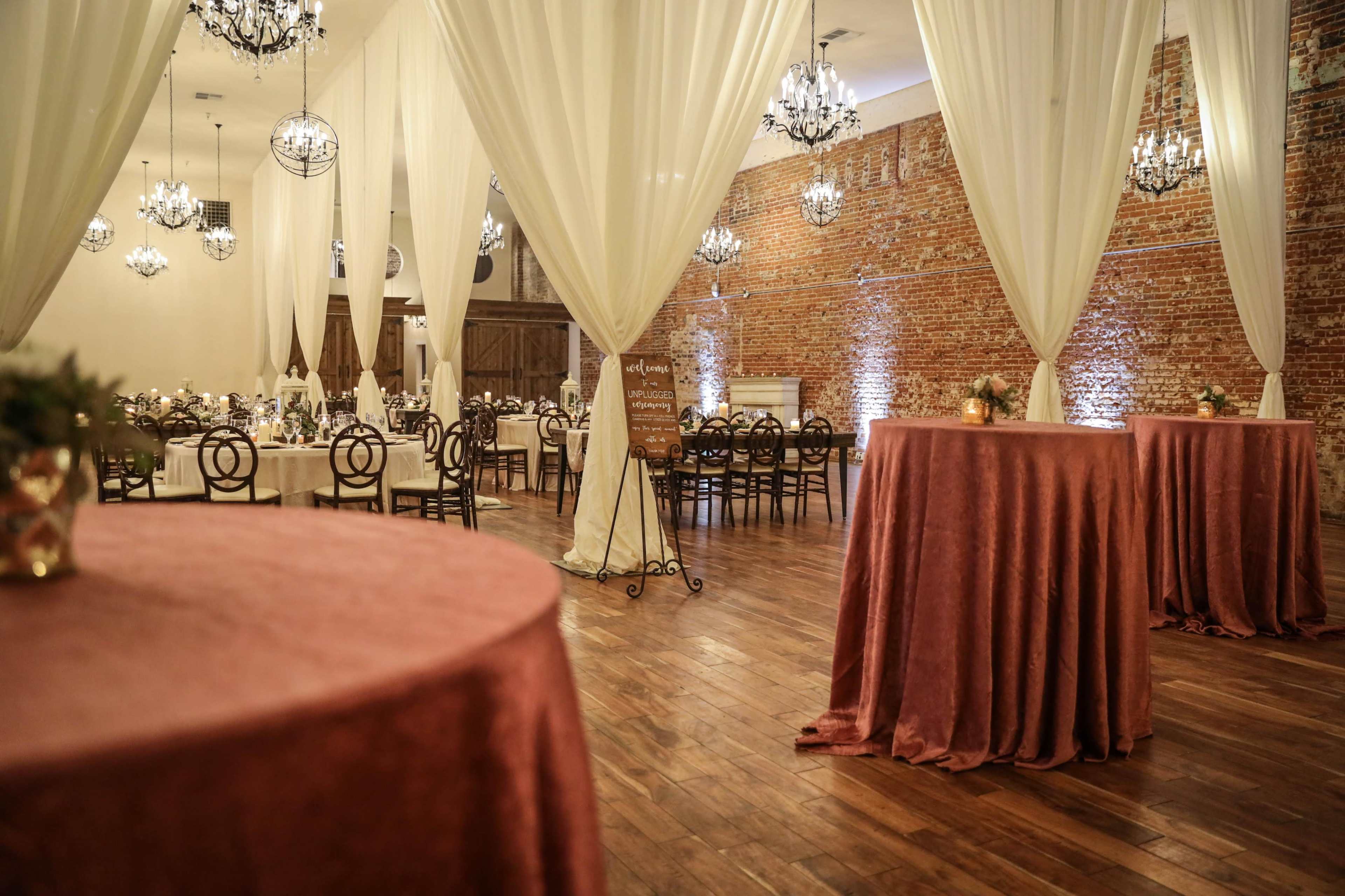 A spacious event hall featuring round tables dressed in rose-colored tablecloths, elegant chandeliers hanging from the ceiling, and exposed brick walls.