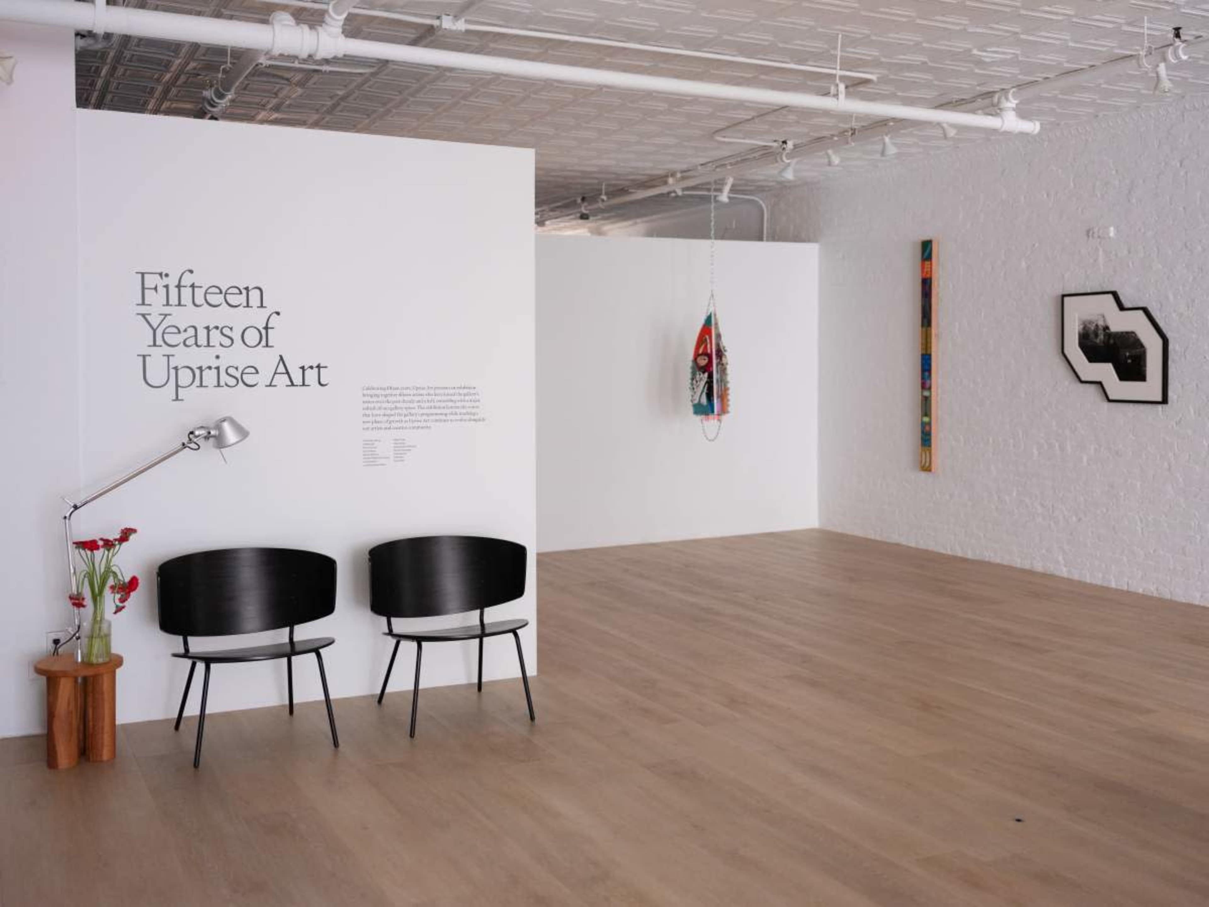 The image shows a spacious art gallery featuring two black chairs, a lamp, a wall with the text "Fifteen Years of Uprise Art," and several artworks hung on the walls.