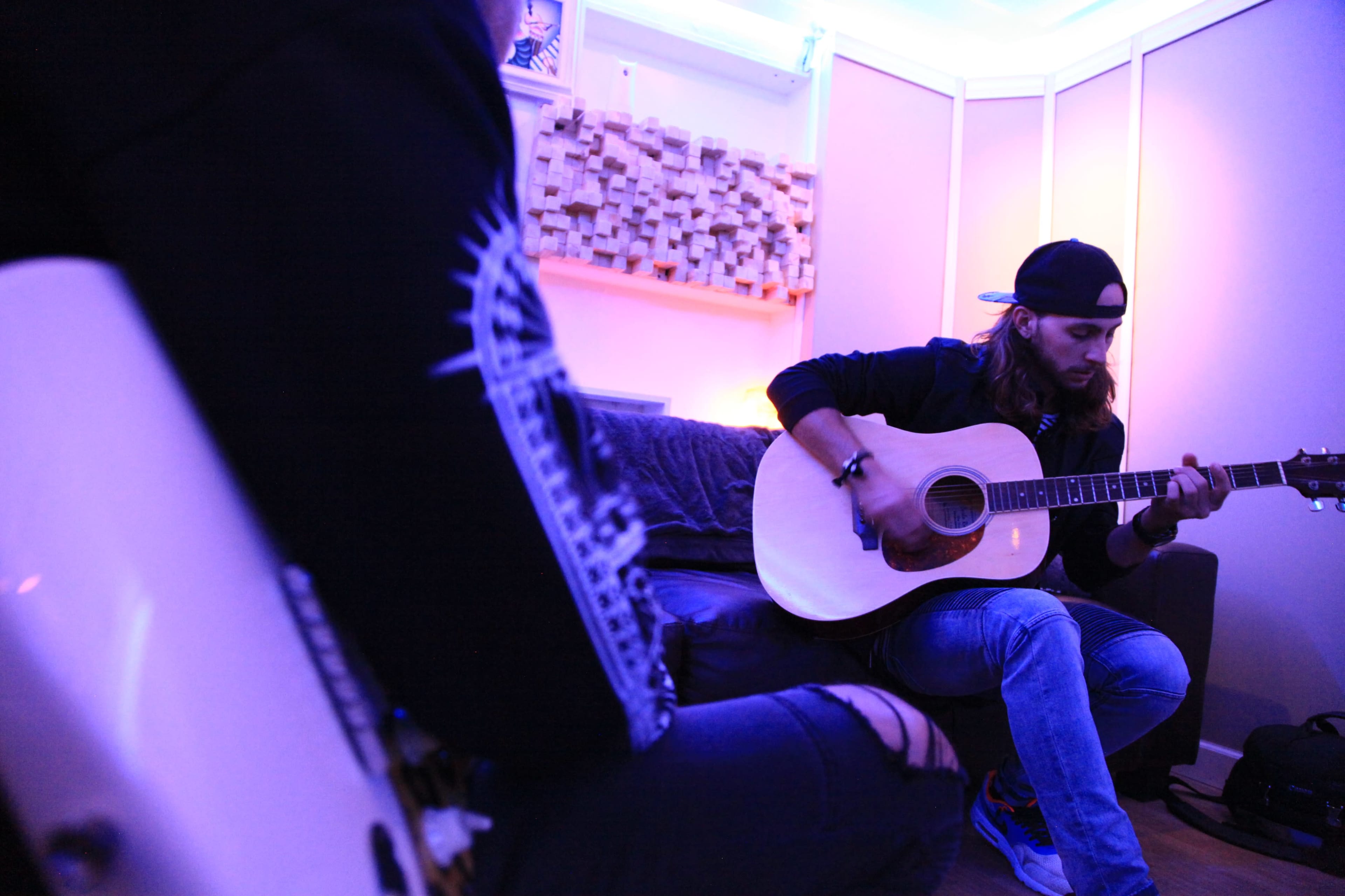 Two musicians are sitting in a room with purple lighting, one playing an acoustic guitar while the other is partially visible with an electric guitar.
