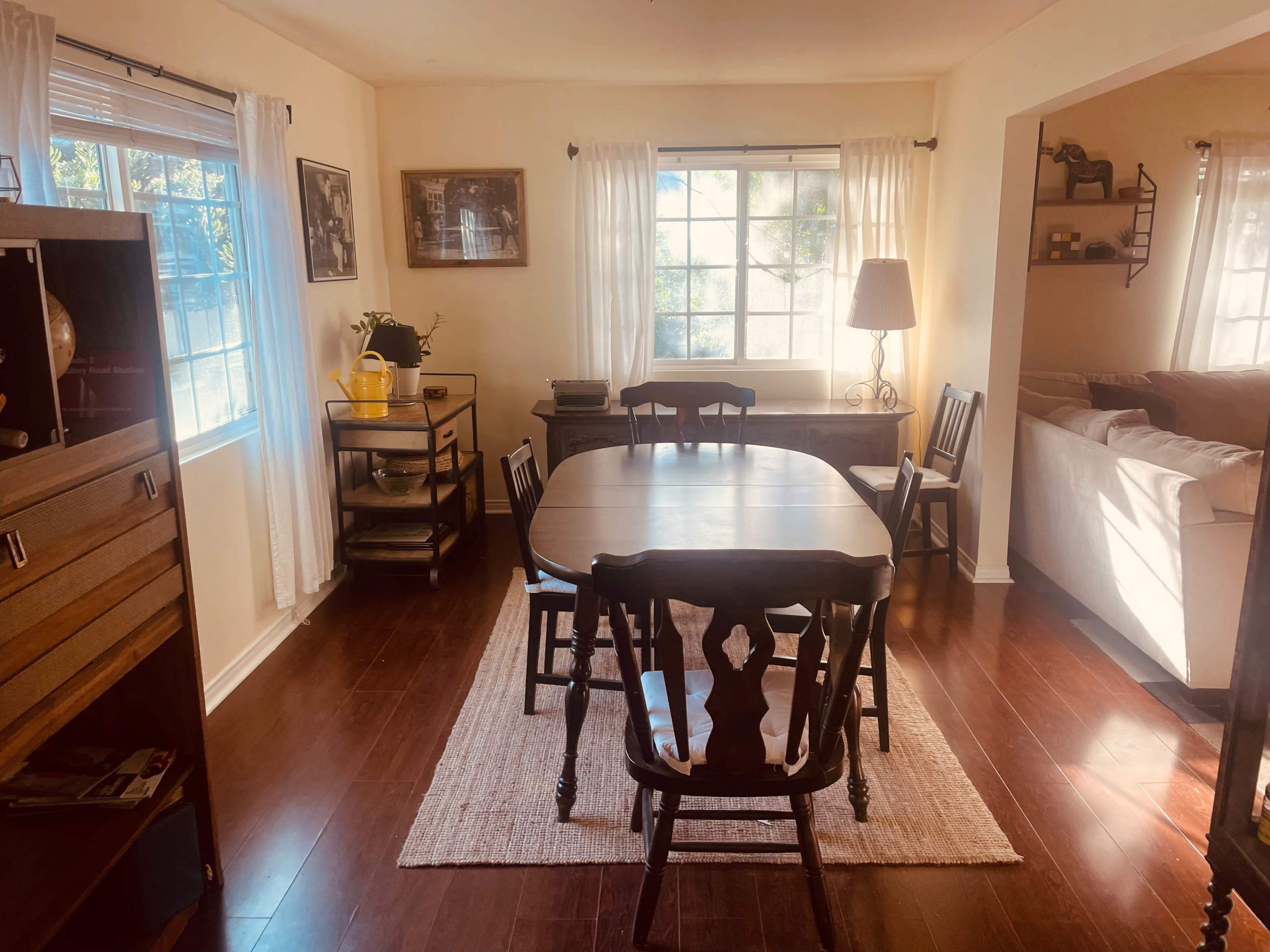 A dining area with a wooden table surrounded by chairs, illuminated by natural light from a large window.