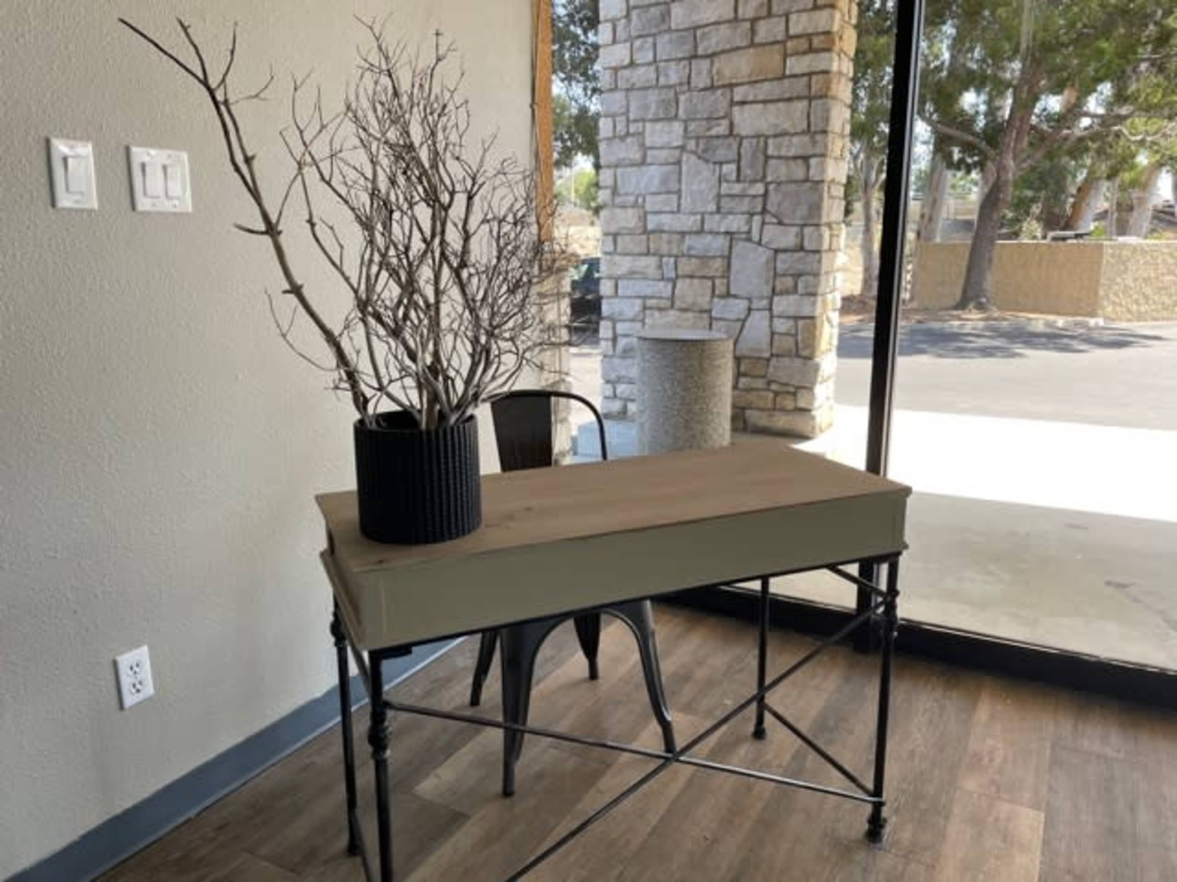 A table with a wooden top and black metal legs holds a black vase with a bare branch next to a window.
