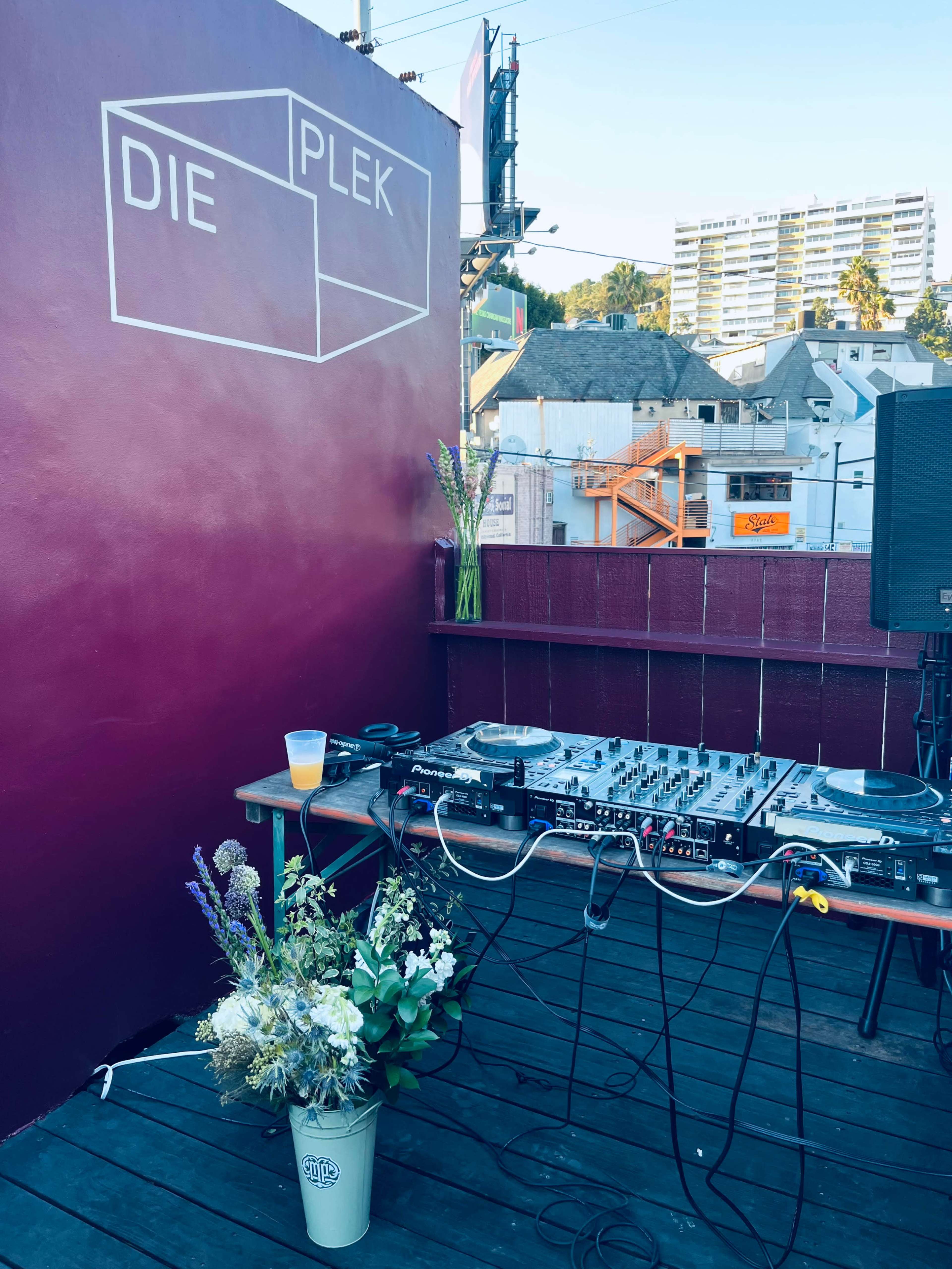 A DJ setup with turntables and a mixer is displayed on a wooden platform, accompanied by a flower arrangement and a drink, against a backdrop of a colorful urban scene.