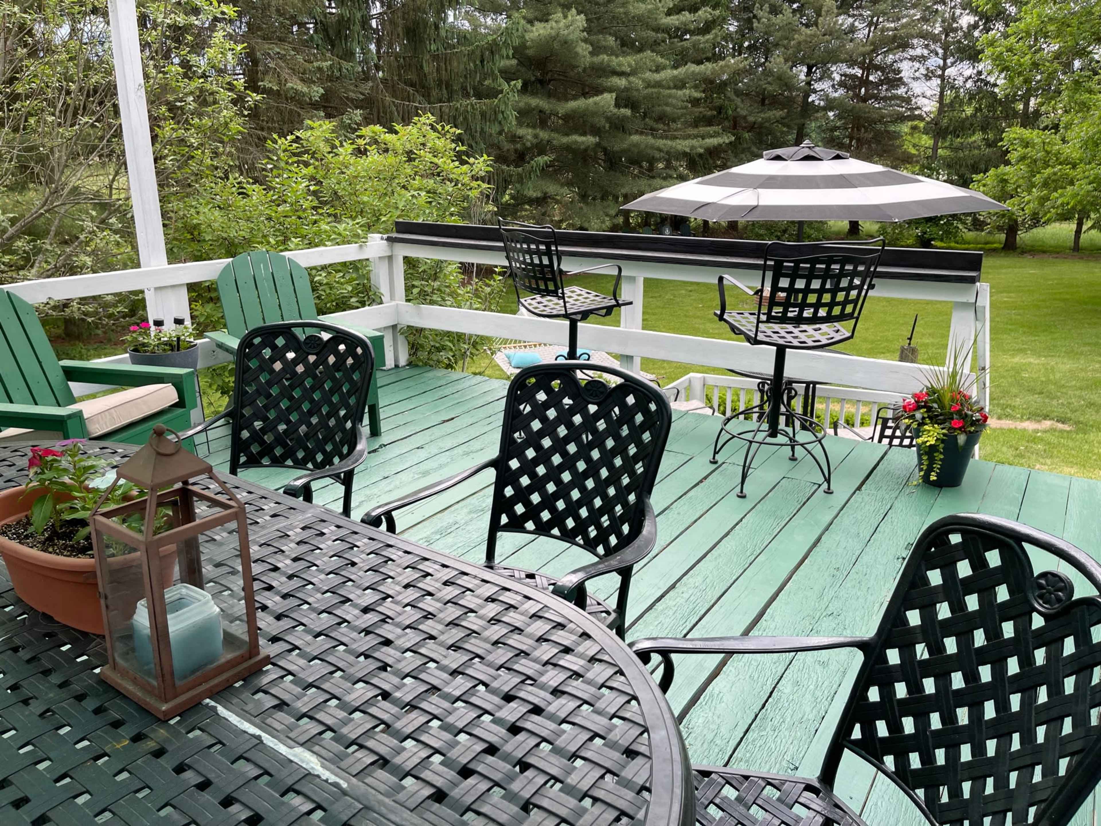 A spacious outdoor deck features black and green chairs, a table with a lantern, and an umbrella over two high-top seats, surrounded by greenery.