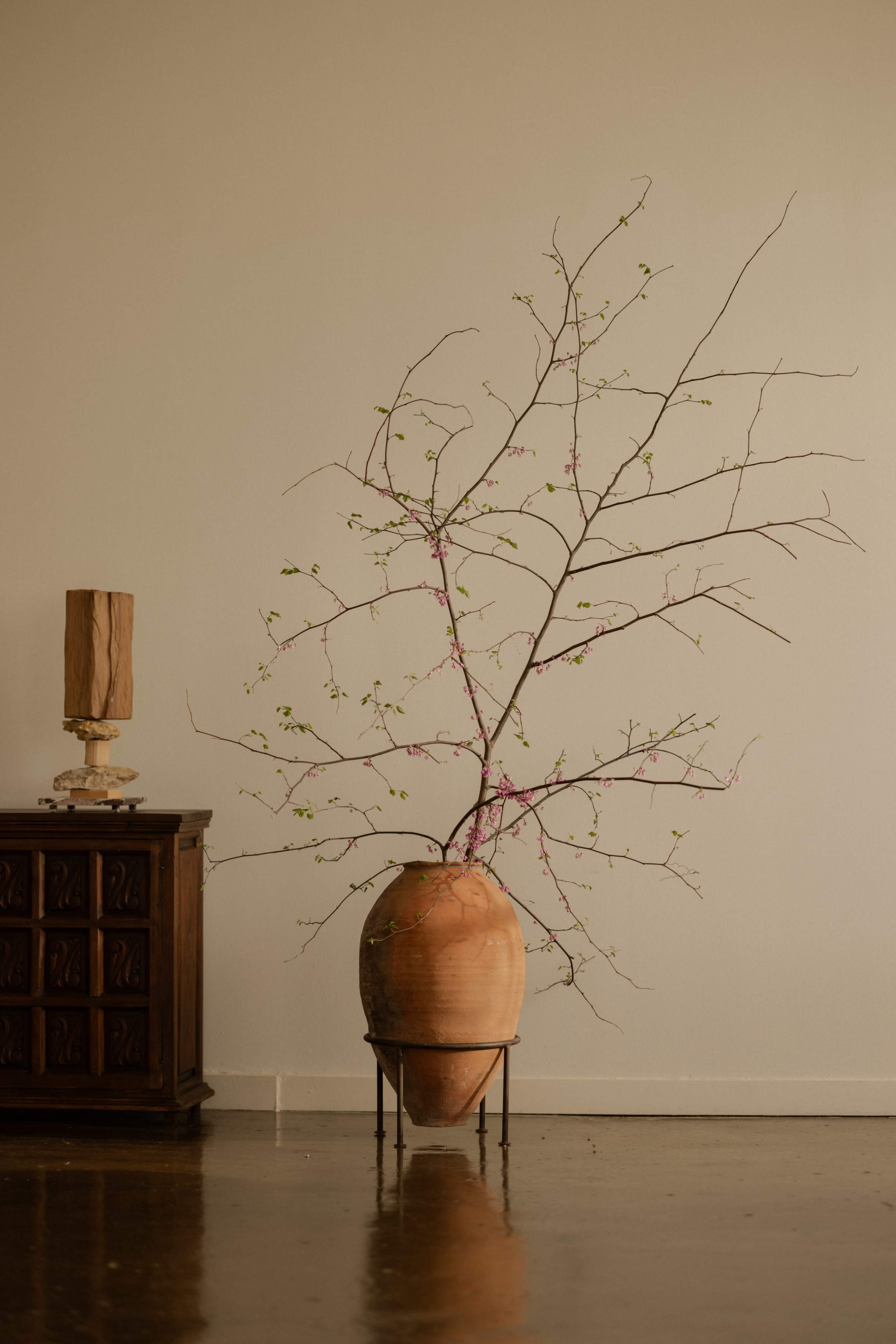 A large terracotta vase stands on a metal stand, containing bare branches with small green leaves and pink flowers, positioned next to a wooden cabinet and lamp in a minimalist room.