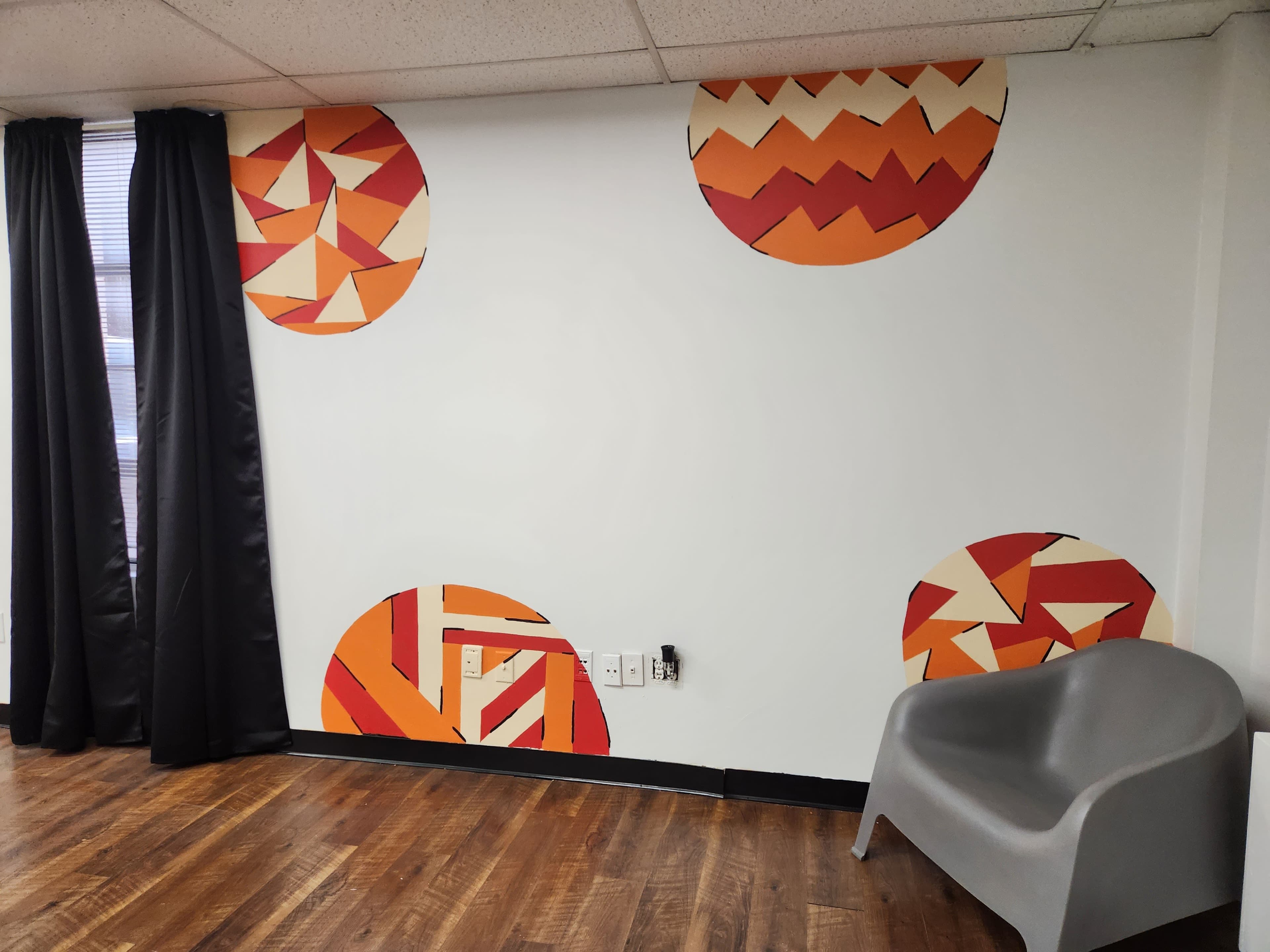 A room with a white wall decorated with geometric orange and red circular patterns, along with dark curtains and a gray chair.