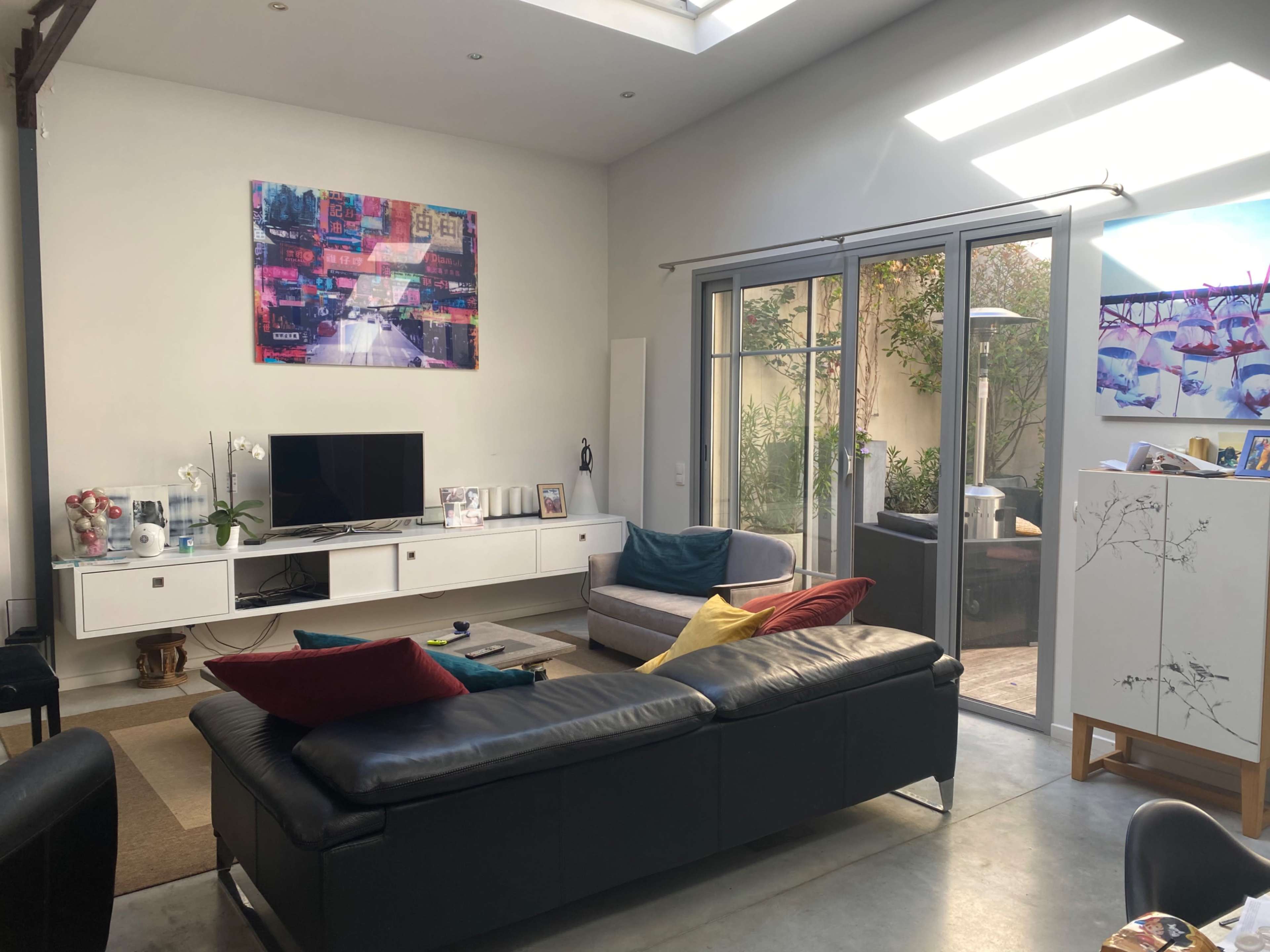 A living room features a sectional couch with colorful throw pillows, a television on a white media console, and large glass doors that open to a garden.