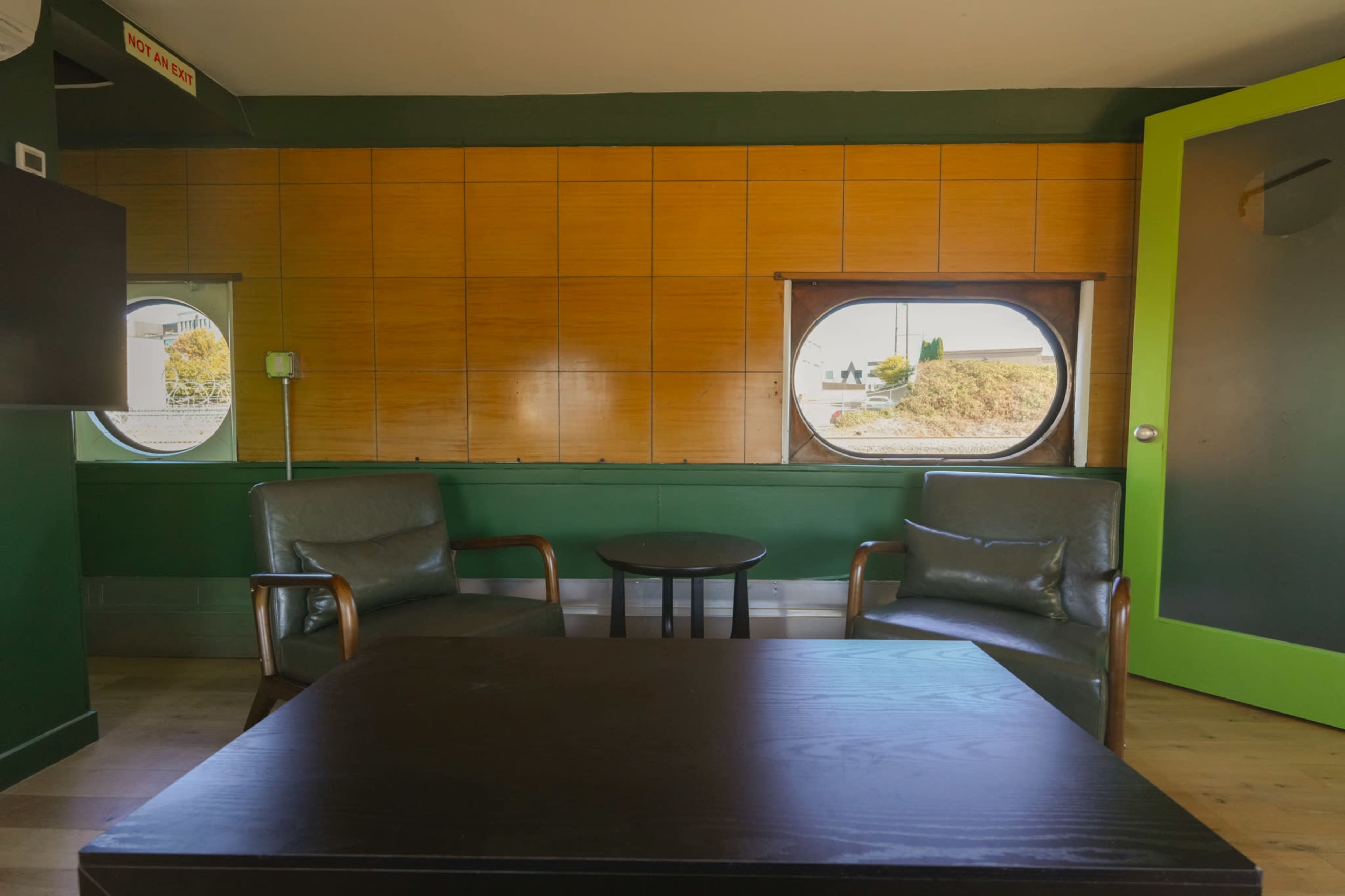 The image shows a minimalist interior space with two green chairs, a round table, and large circular windows surrounded by wooden paneling.