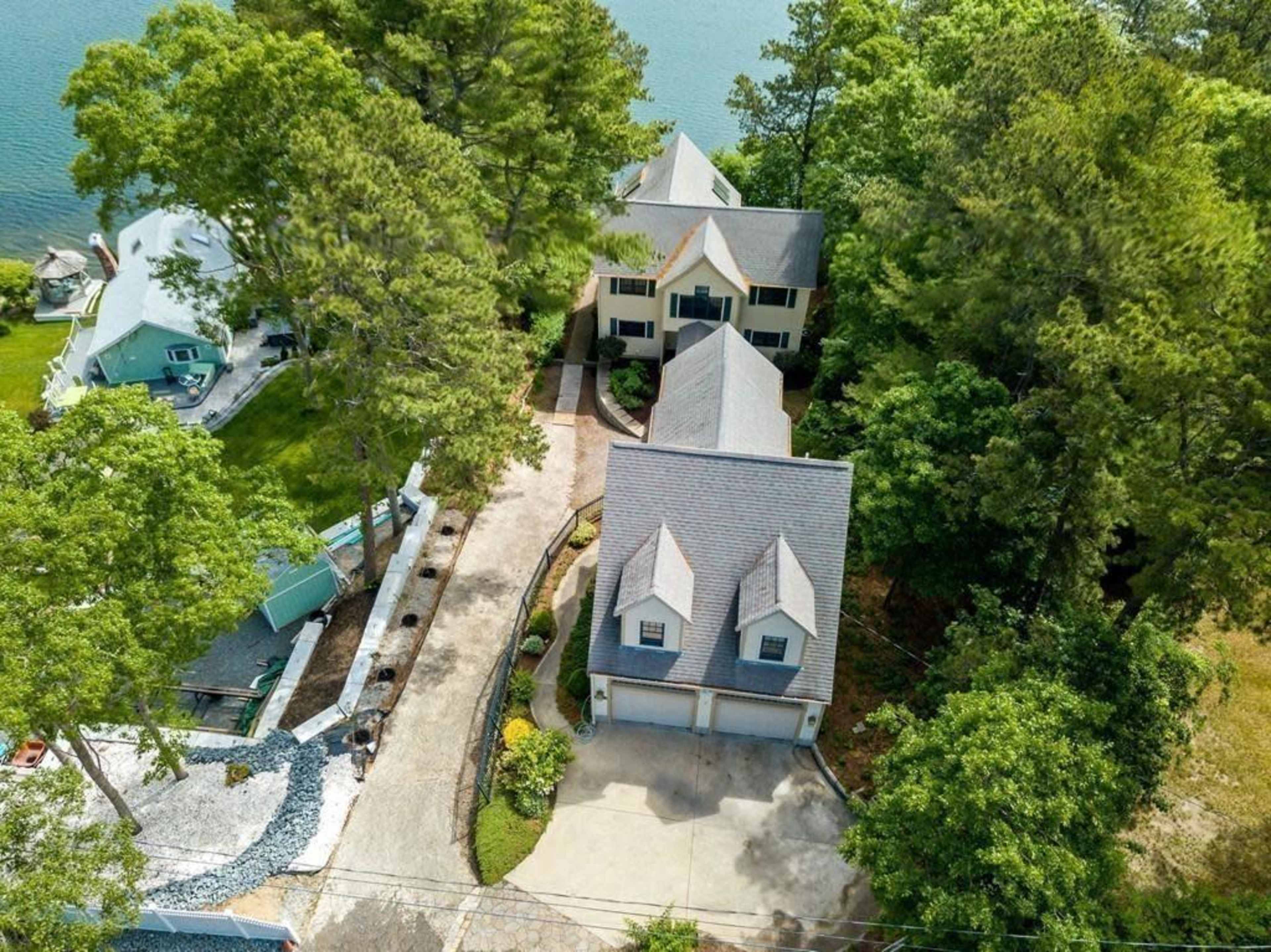 An aerial view shows a large house surrounded by trees with a driveway leading to a two-car garage and a nearby lake.