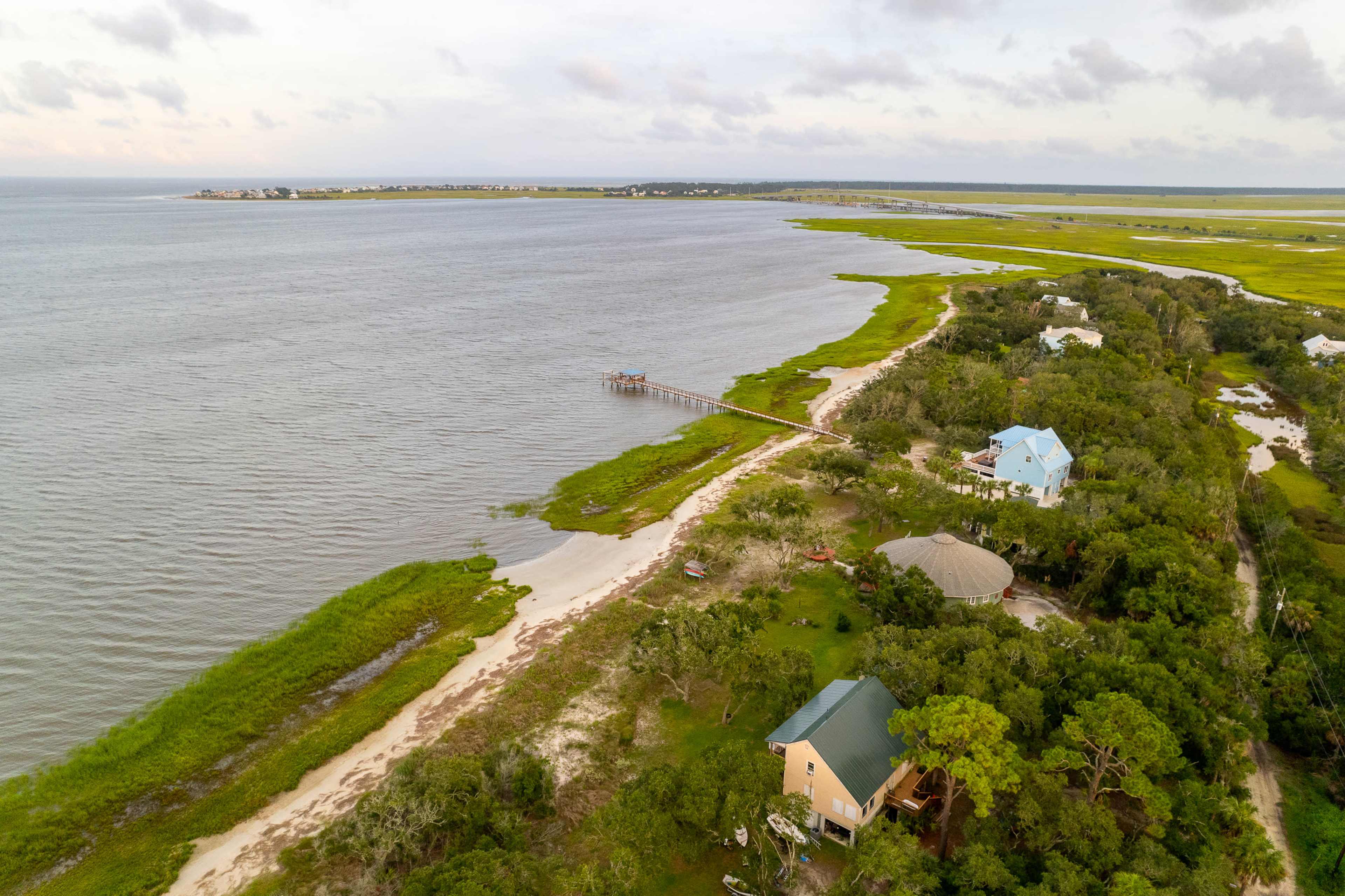 An aerial view shows a coastal landscape with a shallow bay, a wooden pier, and several houses along the shoreline, surrounded by lush greenery.