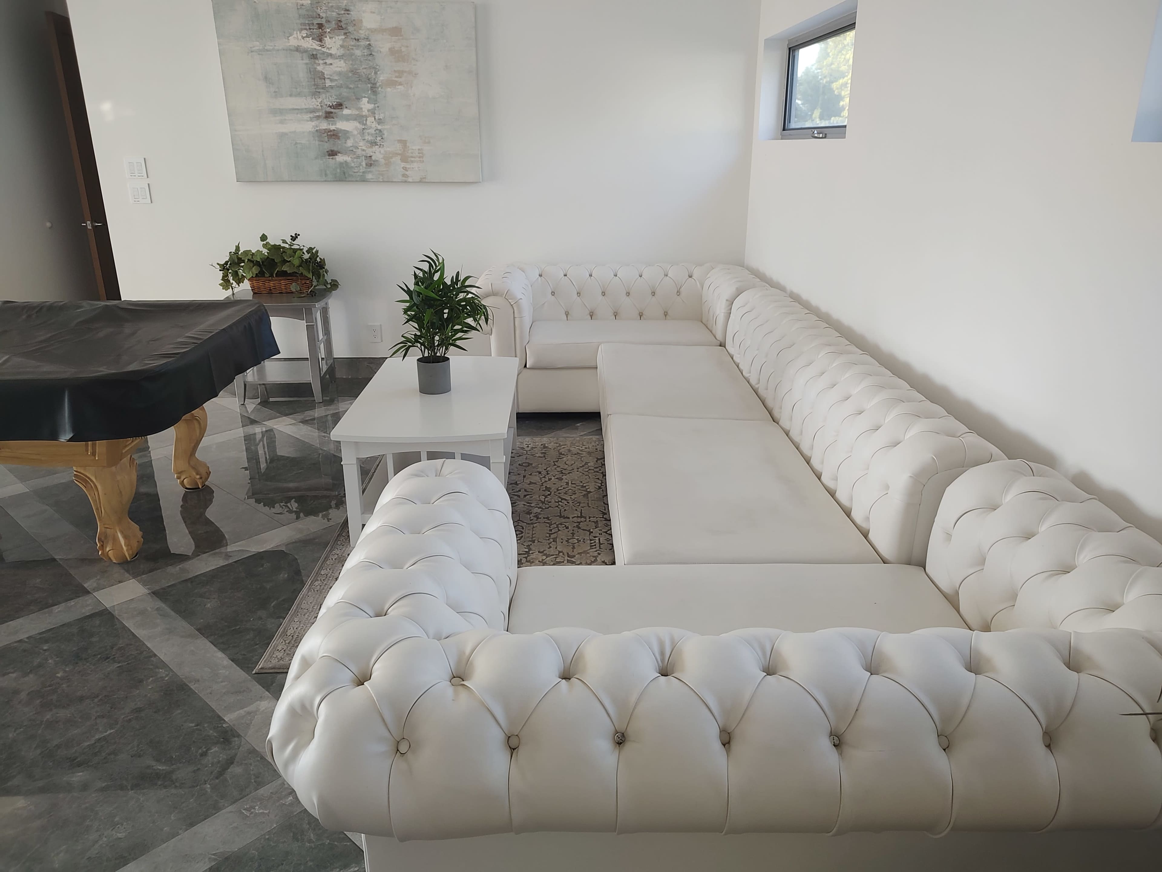 A modern living room featuring a large white tufted sectional sofa, a small white coffee table, and a potted plant, with a pool table in the background.