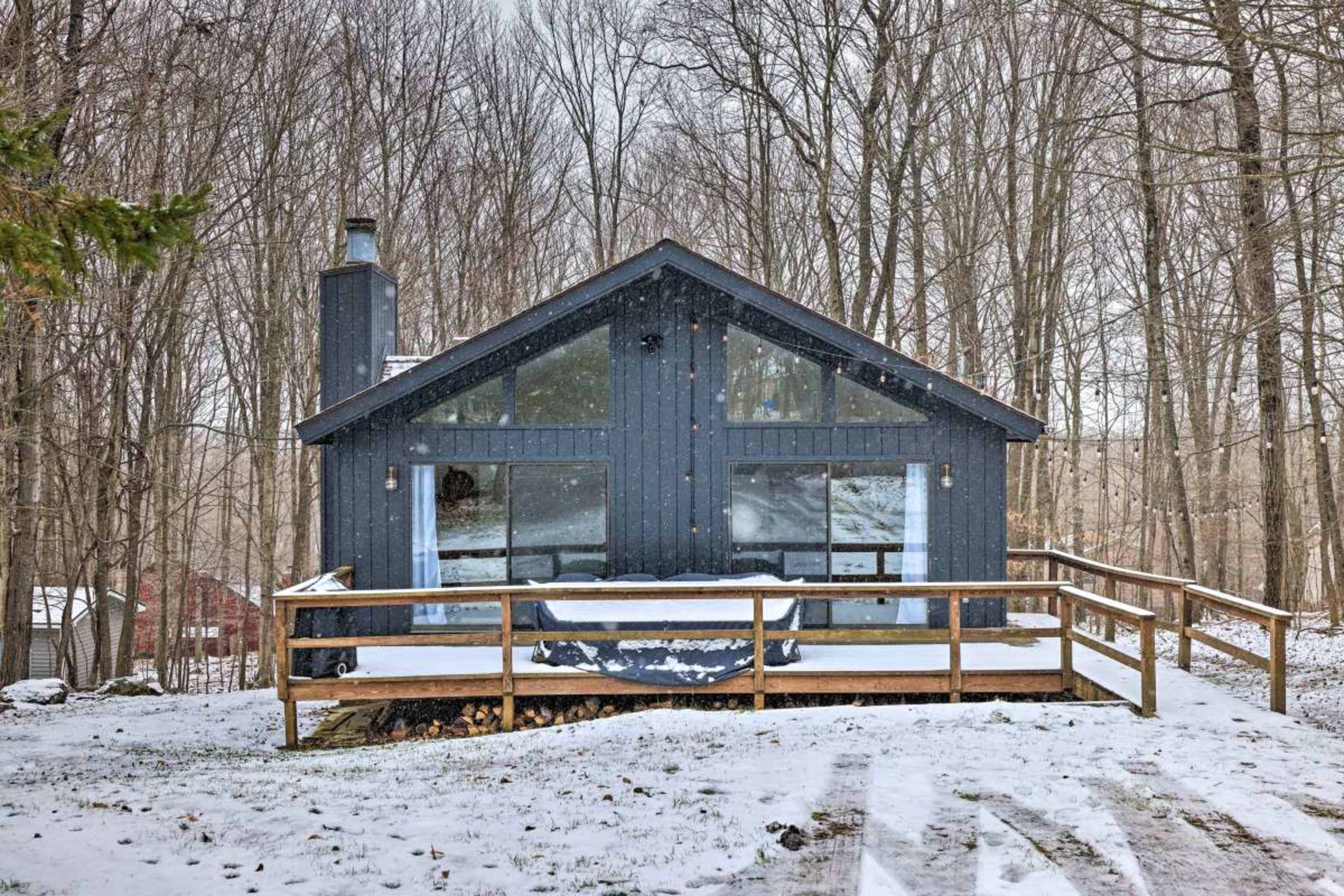 A dark blue cabin with large windows is situated in a snowy forest, featuring a wooden deck and a path leading up to it.