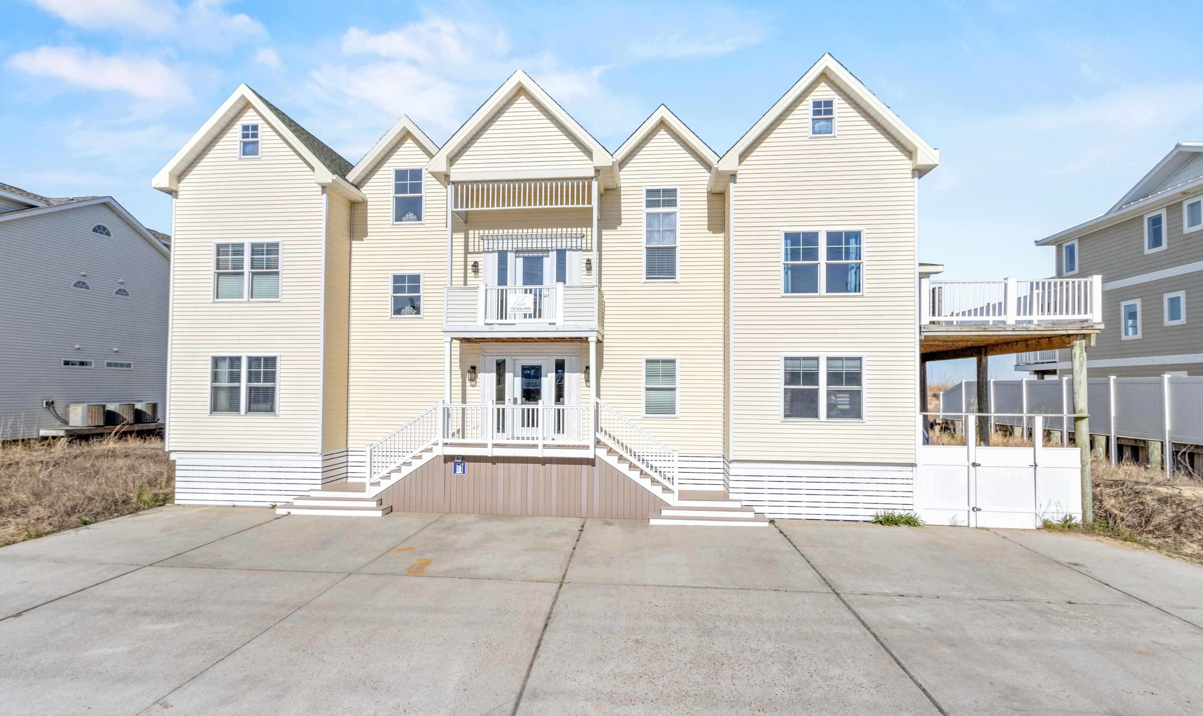 A large, two-story beach house with multiple gabled roofs and a concrete driveway in front, situated in a coastal area with other buildings nearby.