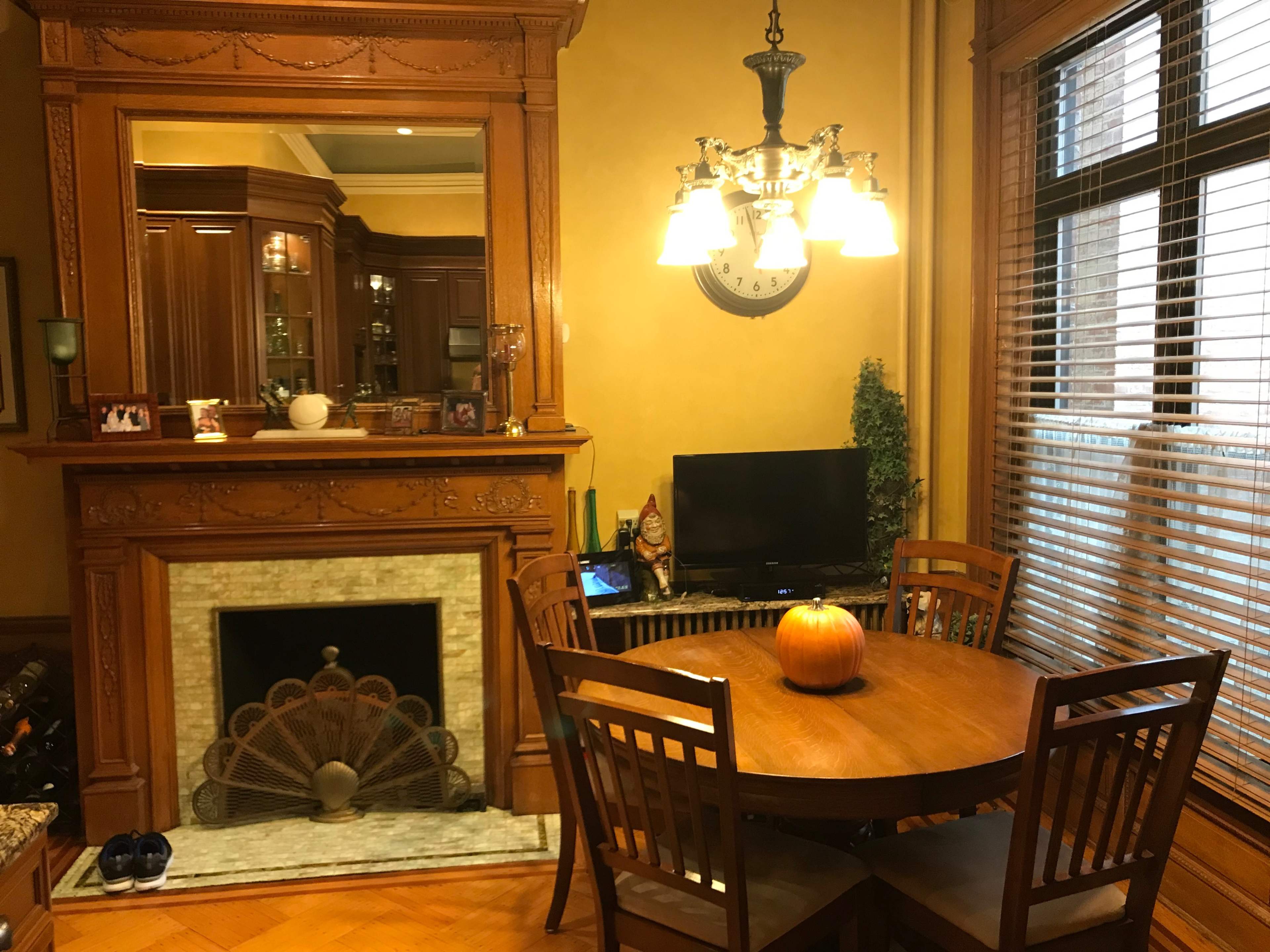 A wooden dining table with four chairs is positioned near a fireplace and a television in a warmly lit room.