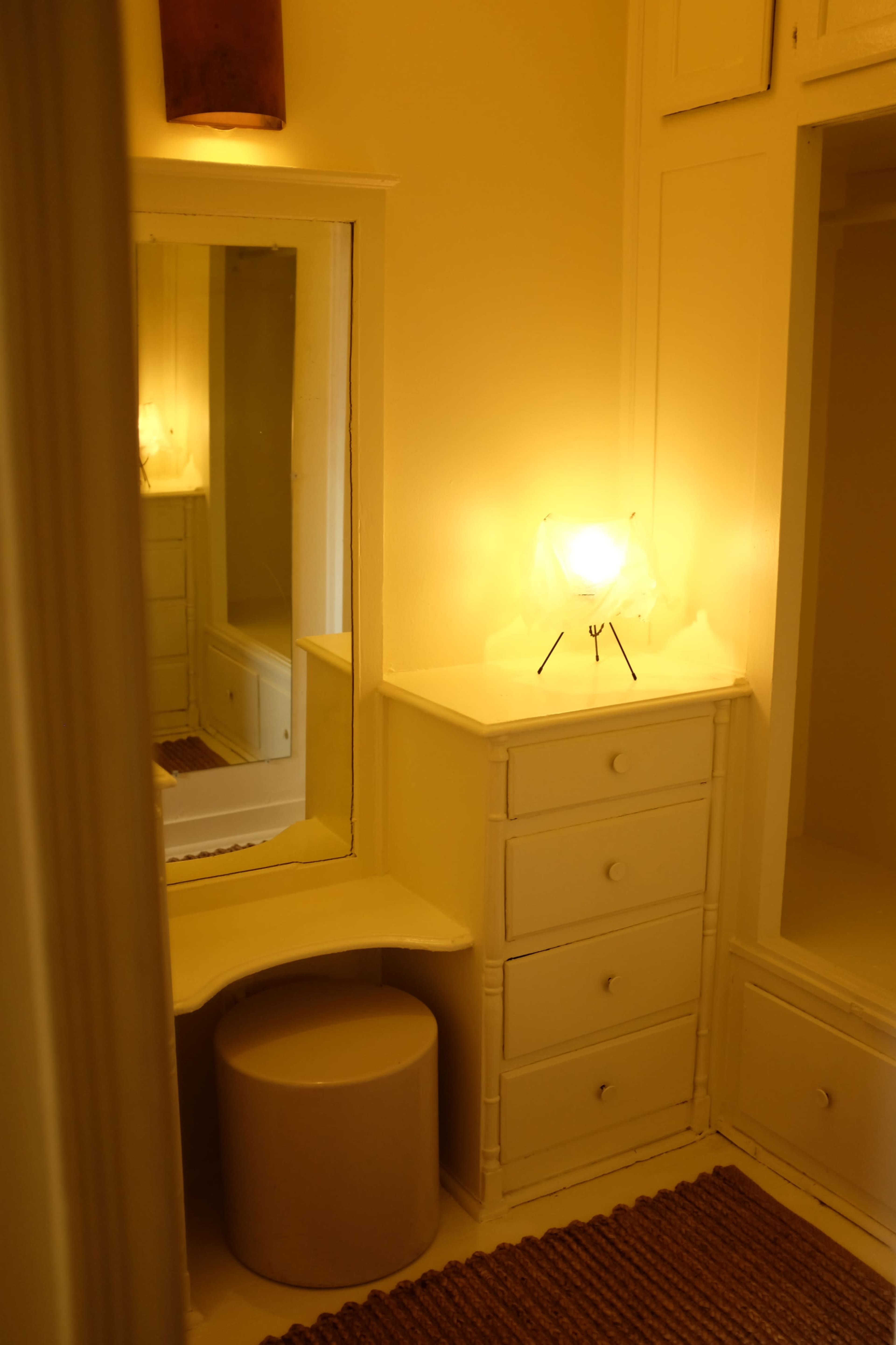 The image shows a small, softly lit dressing area with a mirror, a stool, and a dresser against a bright wall.
